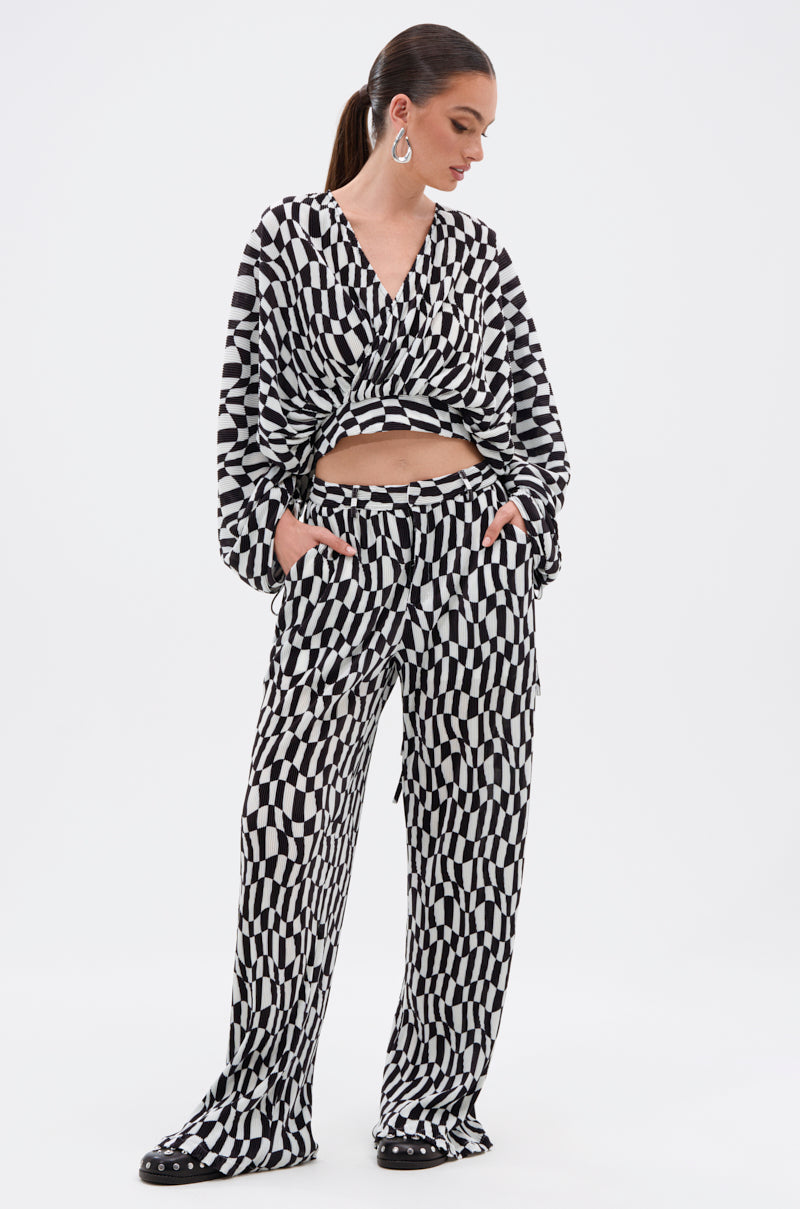 A woman stands against a white background, wearing the BARI CHECKERED TROUSER with a high-rise fit, paired with a long-sleeved black-and-white geometric wrap top, hands in her pockets and looking to the side.