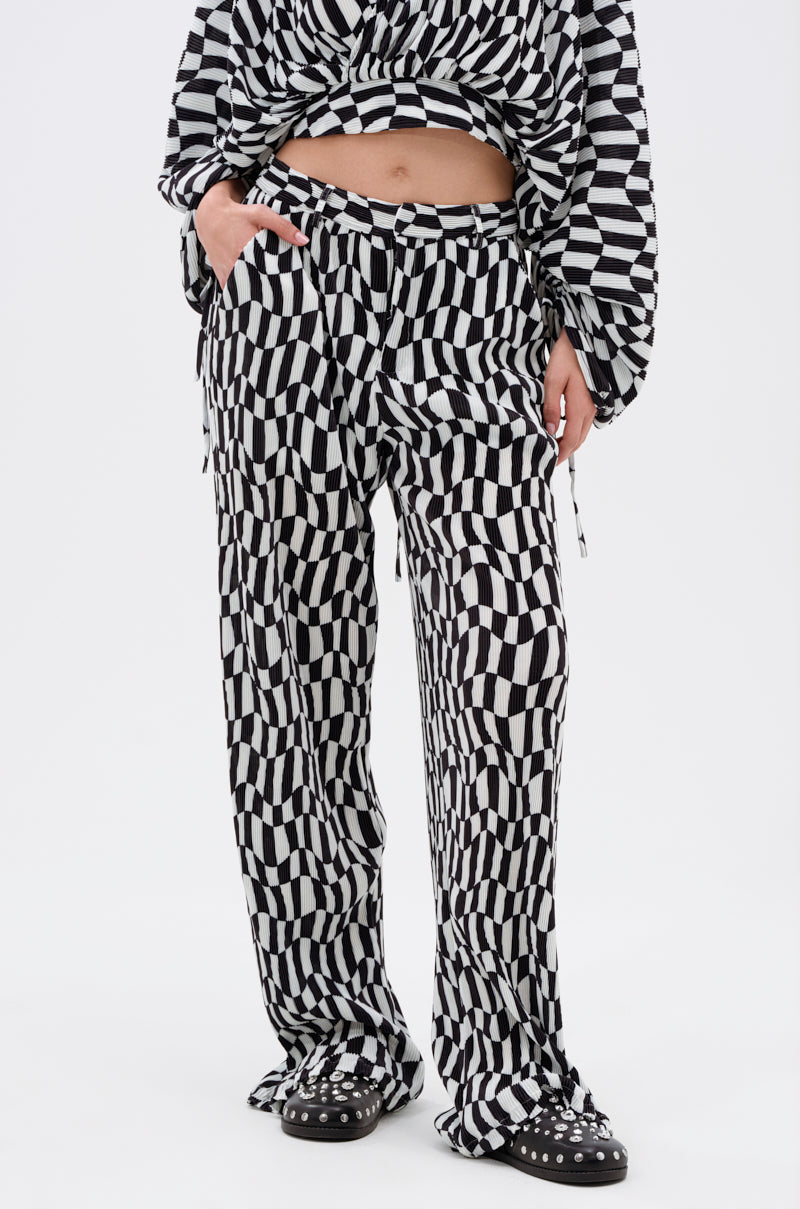 A person wears the BARI CHECKERED TROUSER, featuring a high-rise fit and bold black-and-white geometric patterns. Paired with a matching long-sleeve top, black shoes with white details, one hand in the pocket, against a white background.