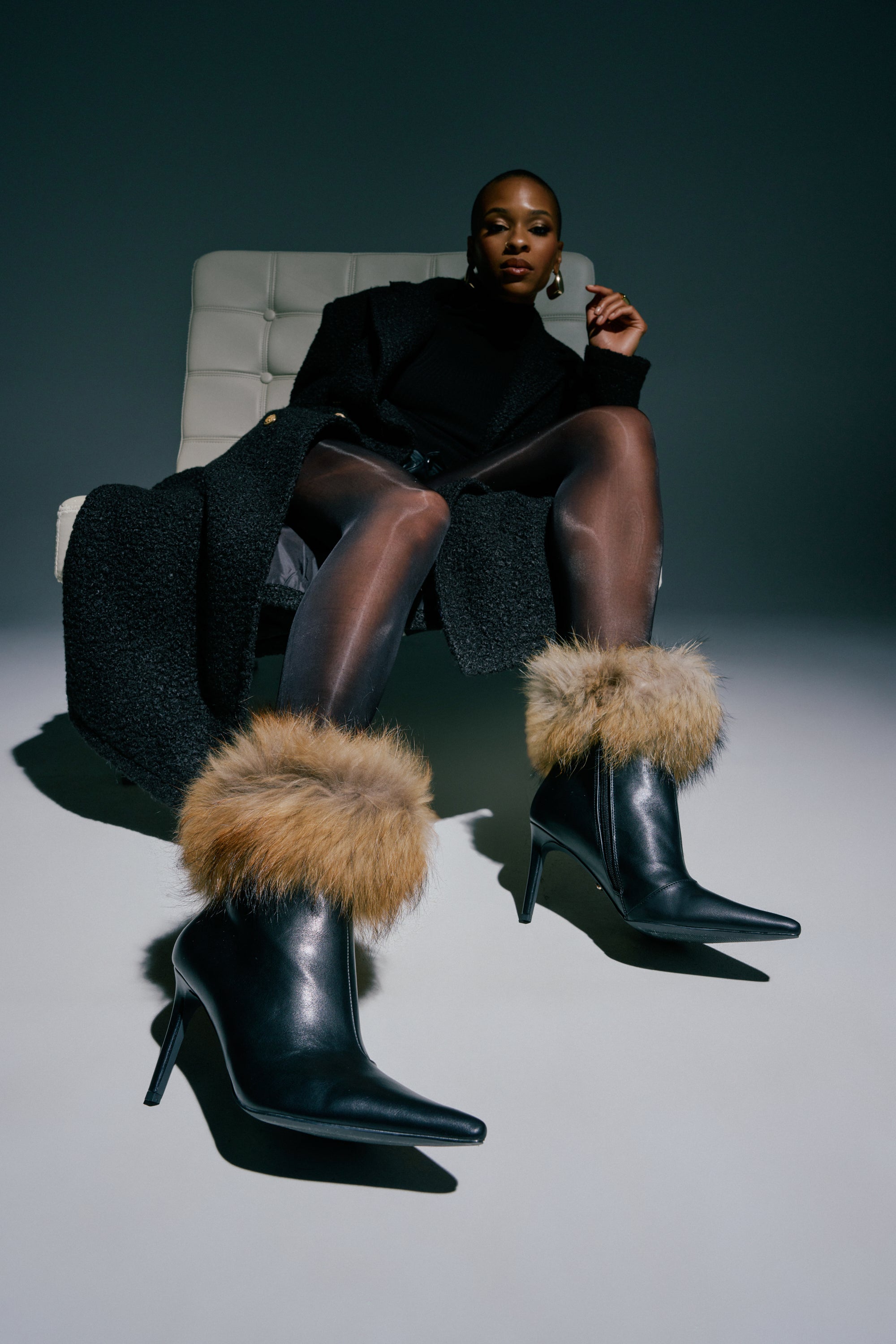 A fashionable individual poses confidently against a dark background, sitting on a modern chair while wearing a black coat, sheer tights, and the AZALEA WANG BARLEY BLACK FUR CUFFED BOOTIE—striking footwear featuring plush fur cuffs and a sleek stiletto heel.