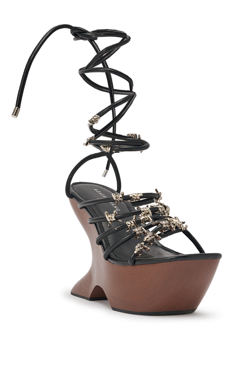 The AKIRA GOLD BARRICADE BLACK SILVER HARDWARE NOVELTY WEDGE SANDAL features black sculptural wedge heels, barbed wire accent straps, silver metal embellishments, a thick brown wooden heel, and long lace-up ankle wraps. 4