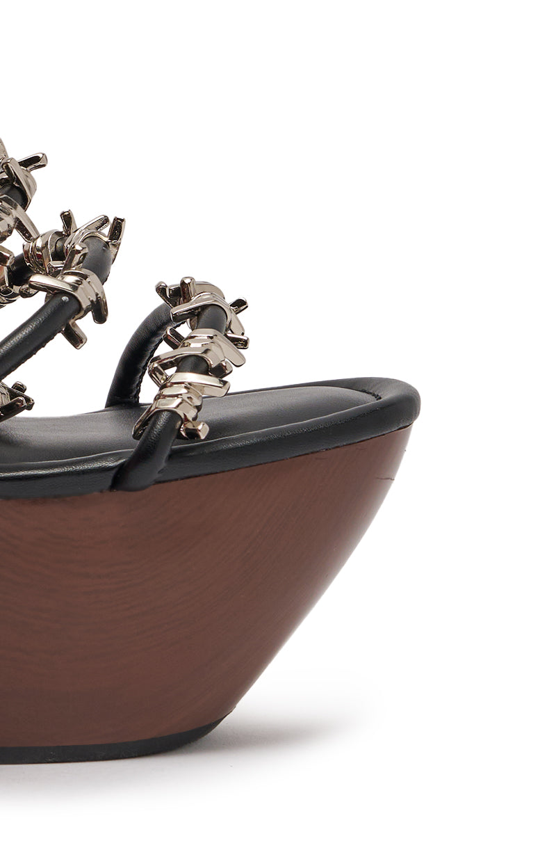 Close-up of the AKIRA GOLD BARRICADE BLACK SILVER HARDWARE NOVELTY WEDGE SANDAL, showcasing black barbed wire-accented straps, metallic details, and a dark wooden sculptural wedge heel against a plain white background. 6