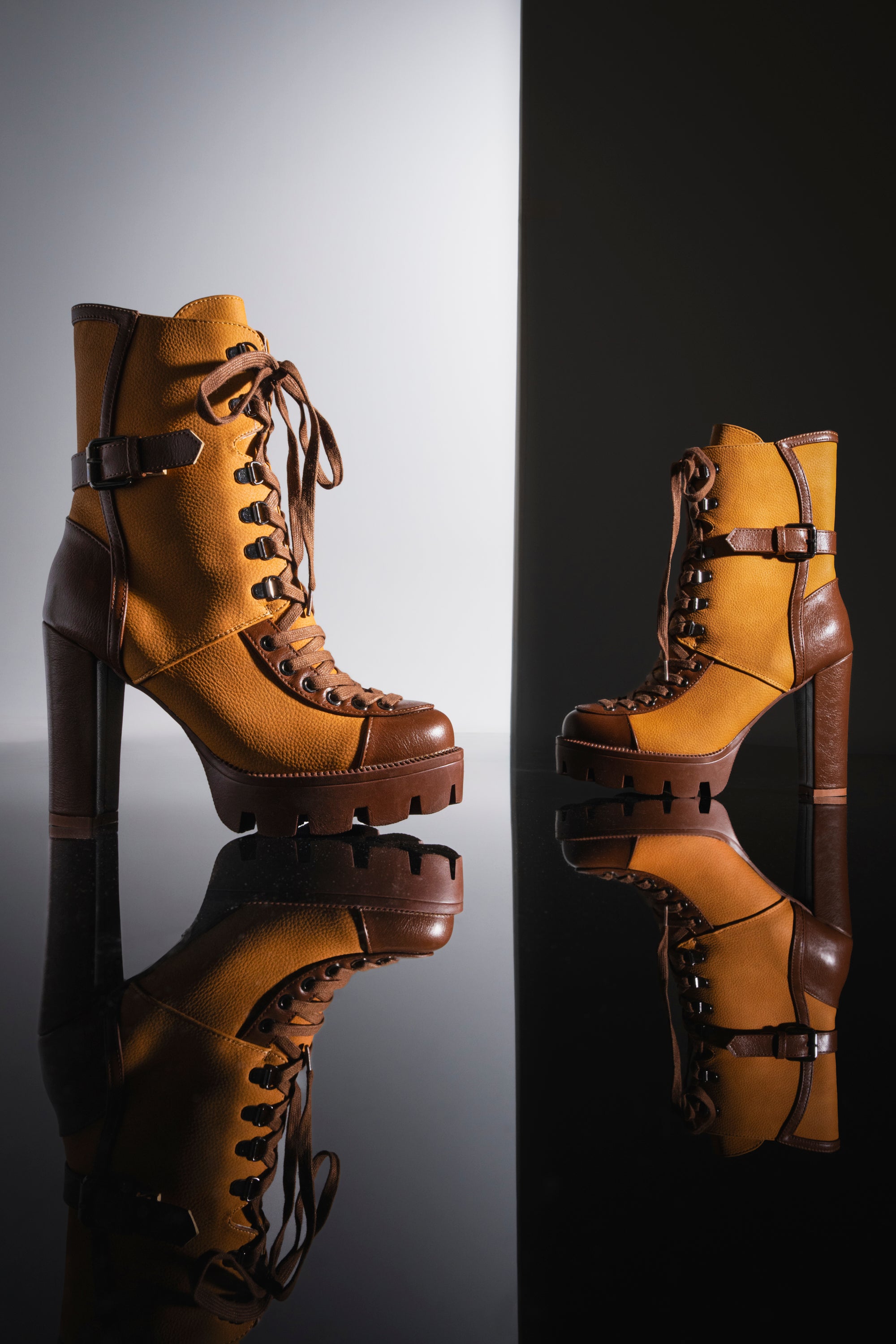 Two AZALEA WANG BARRINGTON Tan Color Blocked Sporty Chunky Booties, featuring a brown and tan design with thick soles and lace-up high heels, stand on a reflective black surface against a light-to-dark gradient background, highlighting their stylish faux leather appeal.
