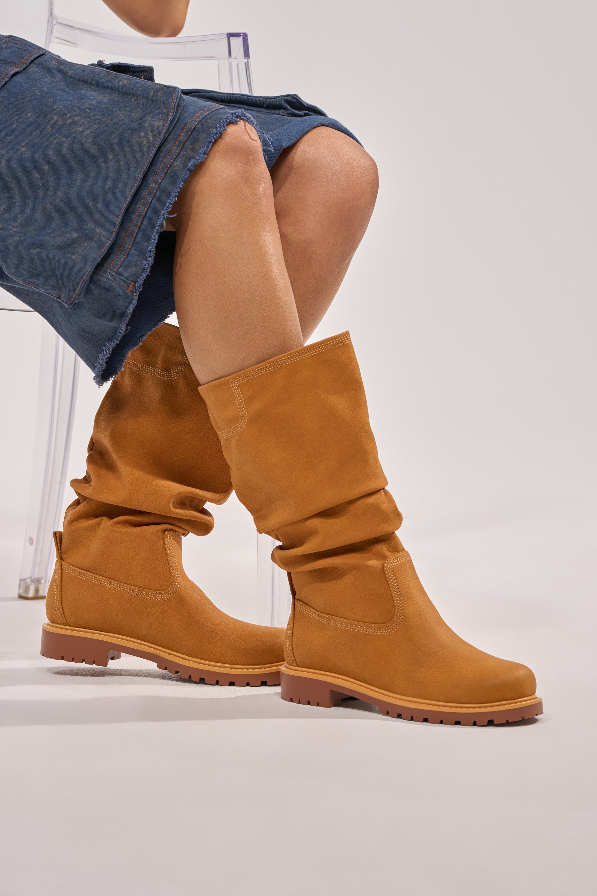 Wearing a frayed denim skirt and AKIRA GOLD BARTLETT TAN FLAT RUCHED BOOTIE, a person sits on a clear chair against a plain light background. 0