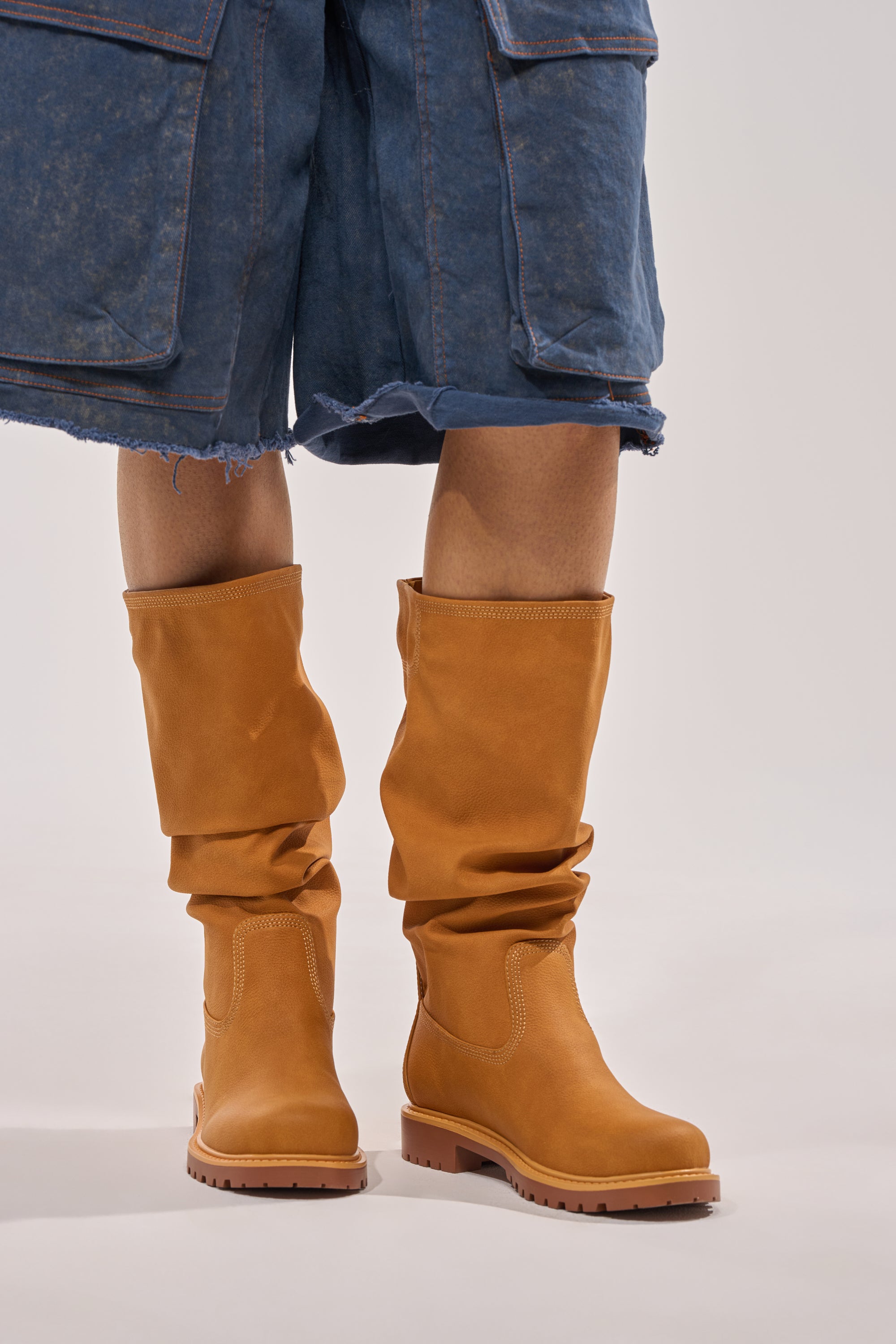 Someone wearing knee-length, frayed blue denim shorts and the AKIRA GOLD BARTLETT TAN FLAT RUCHED BOOTIE stands on a plain light background. 1