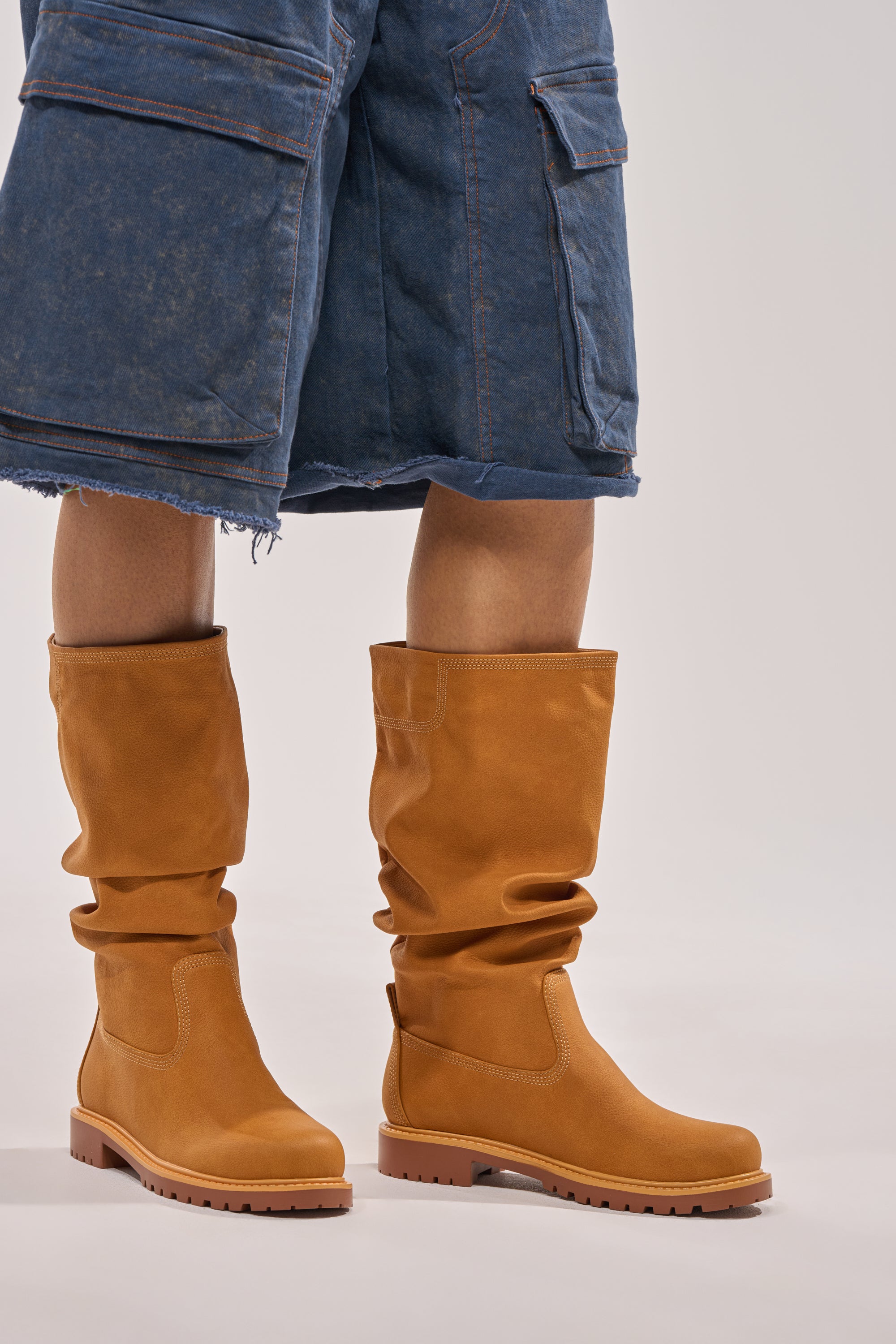 A person wearing the AKIRA GOLD BARTLETT TAN FLAT RUCHED BOOTIE and knee-length blue denim cargo shorts with frayed hems stands against a light background. The booties are tan, ruched, and flat. 2