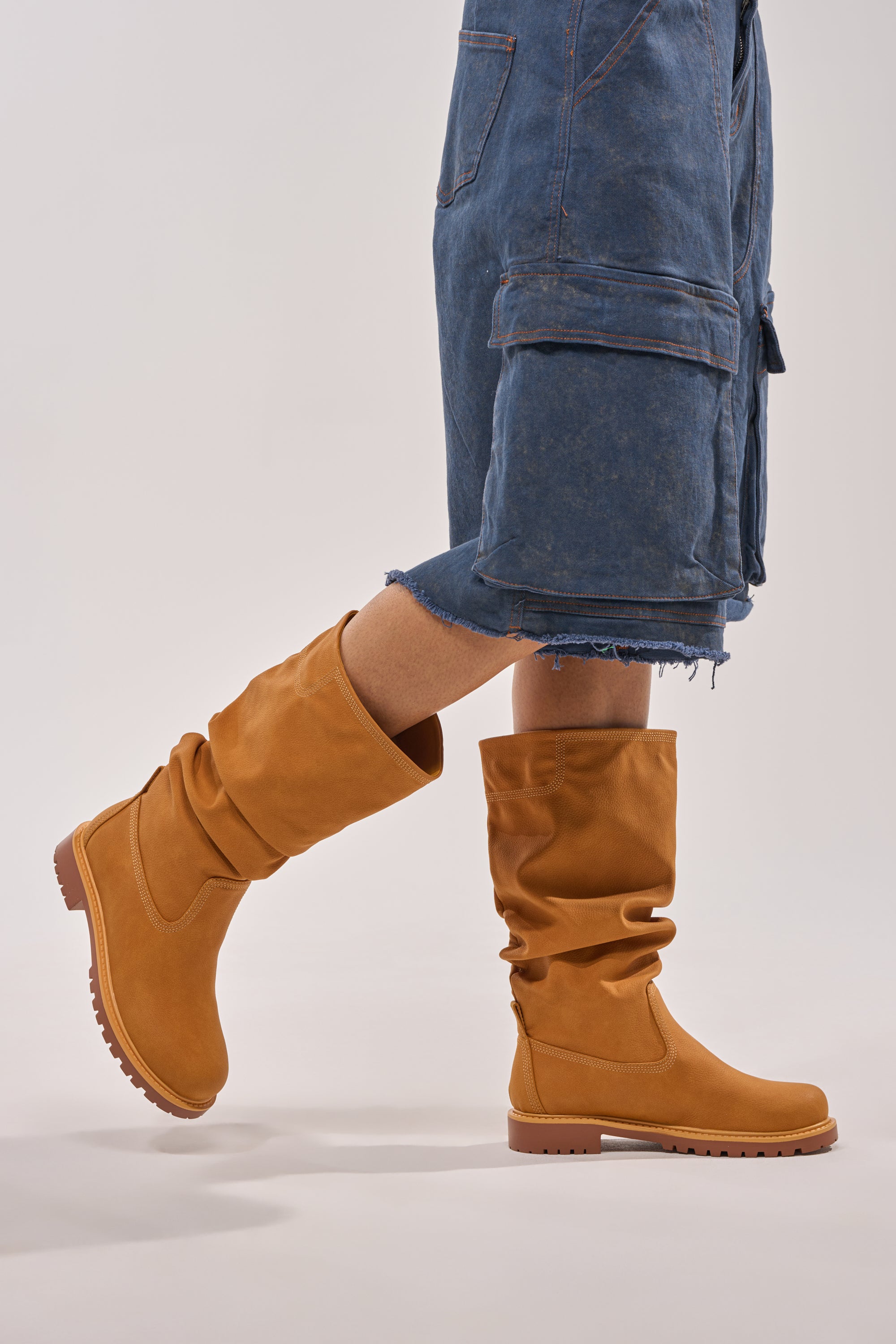 A person wears AKIRA GOLD BARTLETT TAN FLAT RUCHED BOOTIE boots and blue denim shorts, standing against a light background with only the lower half of the body visible. 3