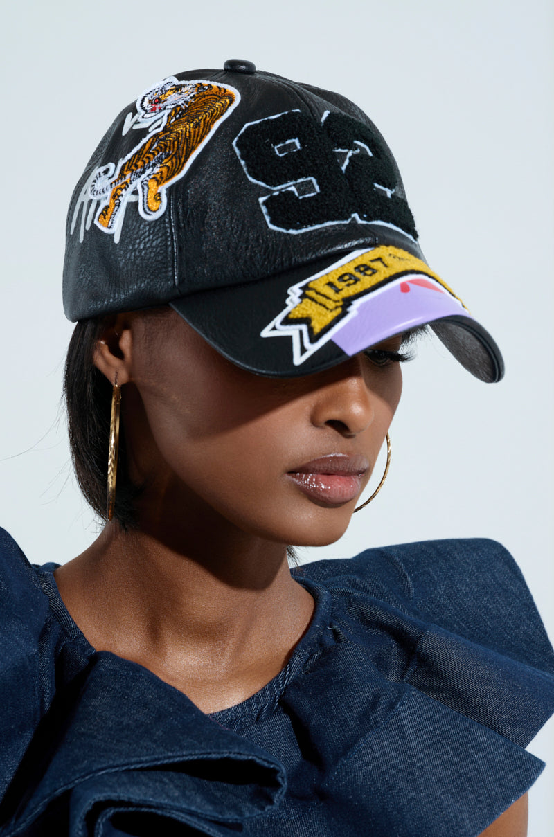 A person wearing the REALLY LIKE THAT DETAIL HAT, featuring letterman-style patches such as a tiger, the number 93, and the year 1991. They complement the look with gold hoop earrings and a dark blue textured outfit.