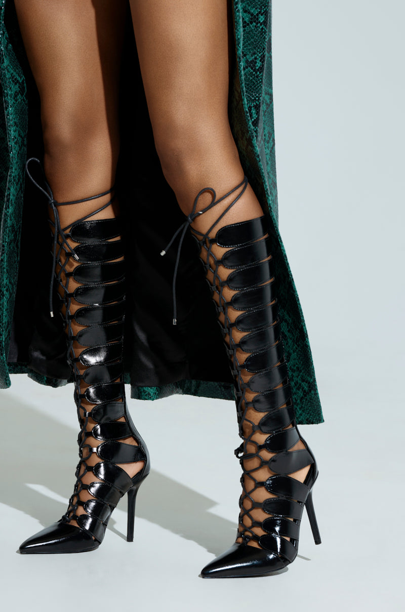 A person struts in AZALEA WANG BASTIAN BLACK BOOT, featuring glossy vegan leather, knee-high lace-ups, pointed toes, and thin high heels with cutout designs, paired with a dark green textured skirt.