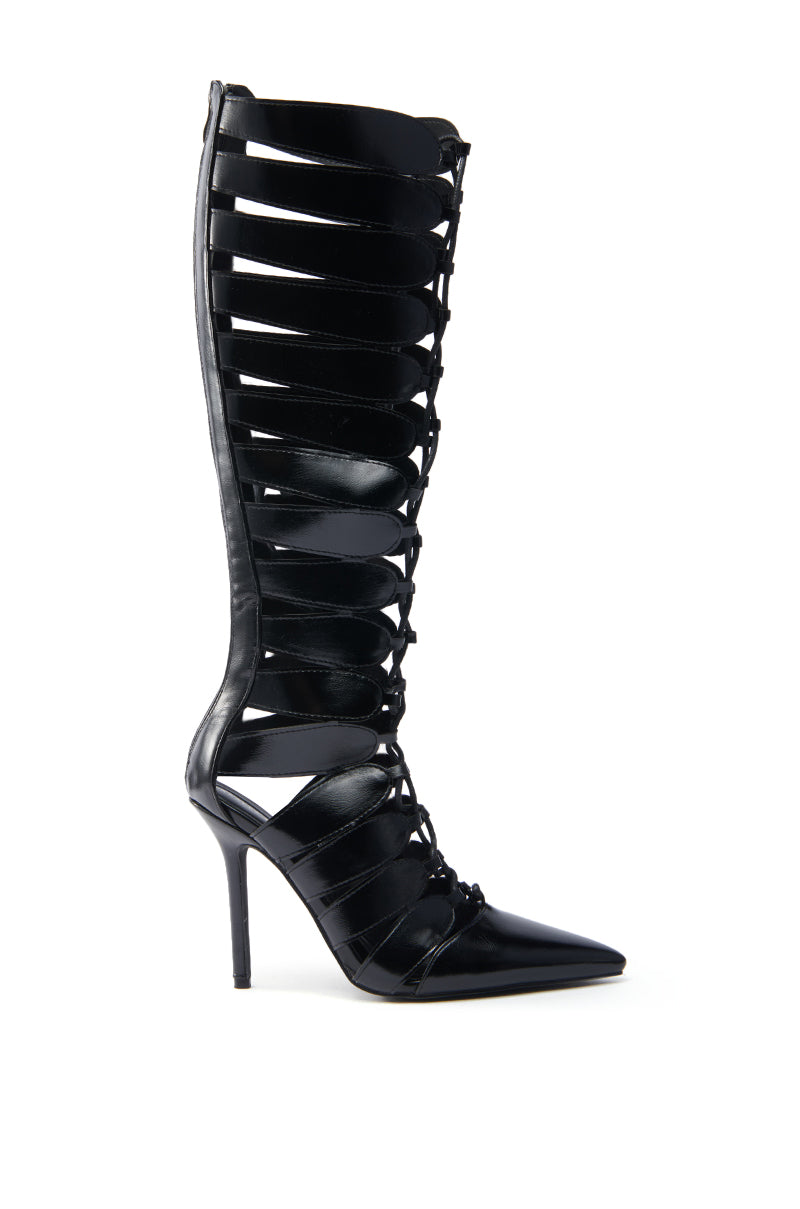 The AZALEA WANG BASTIAN BLACK BOOT is a single black high-heeled gladiator boot crafted from vegan leather, featuring a pointed toe, multiple horizontal straps, an open calf design fastened with laces, and a thin stiletto-style heel.