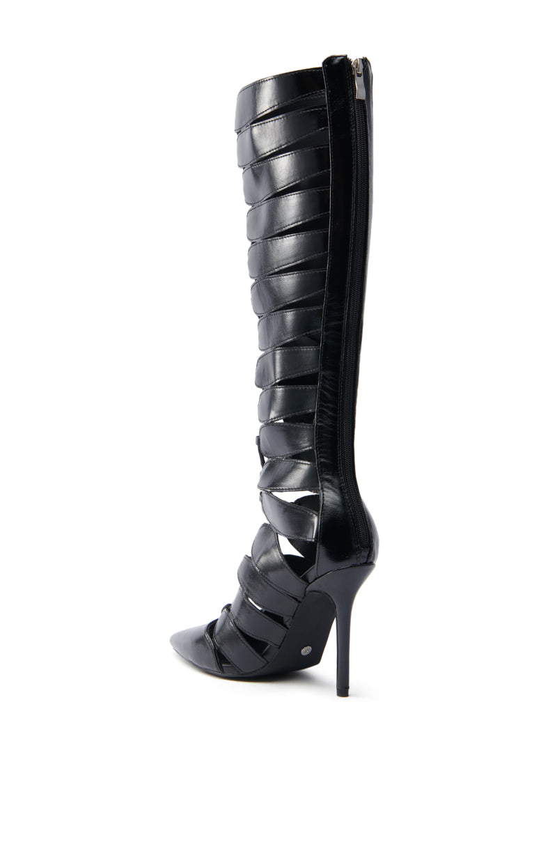 The AZALEA WANG BASTIAN BLACK BOOT is a stylish vegan leather knee-high boot with a pointed toe, unique strappy design, and slender stiletto heel. It features a zipper closure at the back for an elegant look.