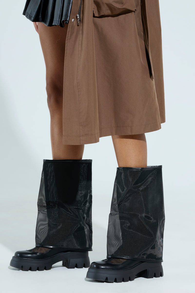 A person in a brown garment is shown with visible lower legs, wearing distinctive AZALEA WANG Baye Black Mesh Boots, known for their glossy, chunky soles that rise to mid-calf. The backdrop is plain light gray.
