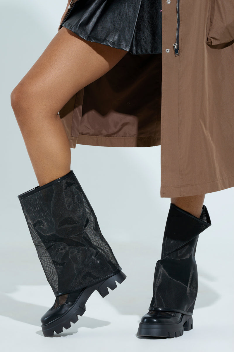 A person is wearing a brown coat, a short black leather skirt, and the AZALEA WANG BAYE BLACK MESH BOOT with mid-calf height and mesh details, all set against a light background.