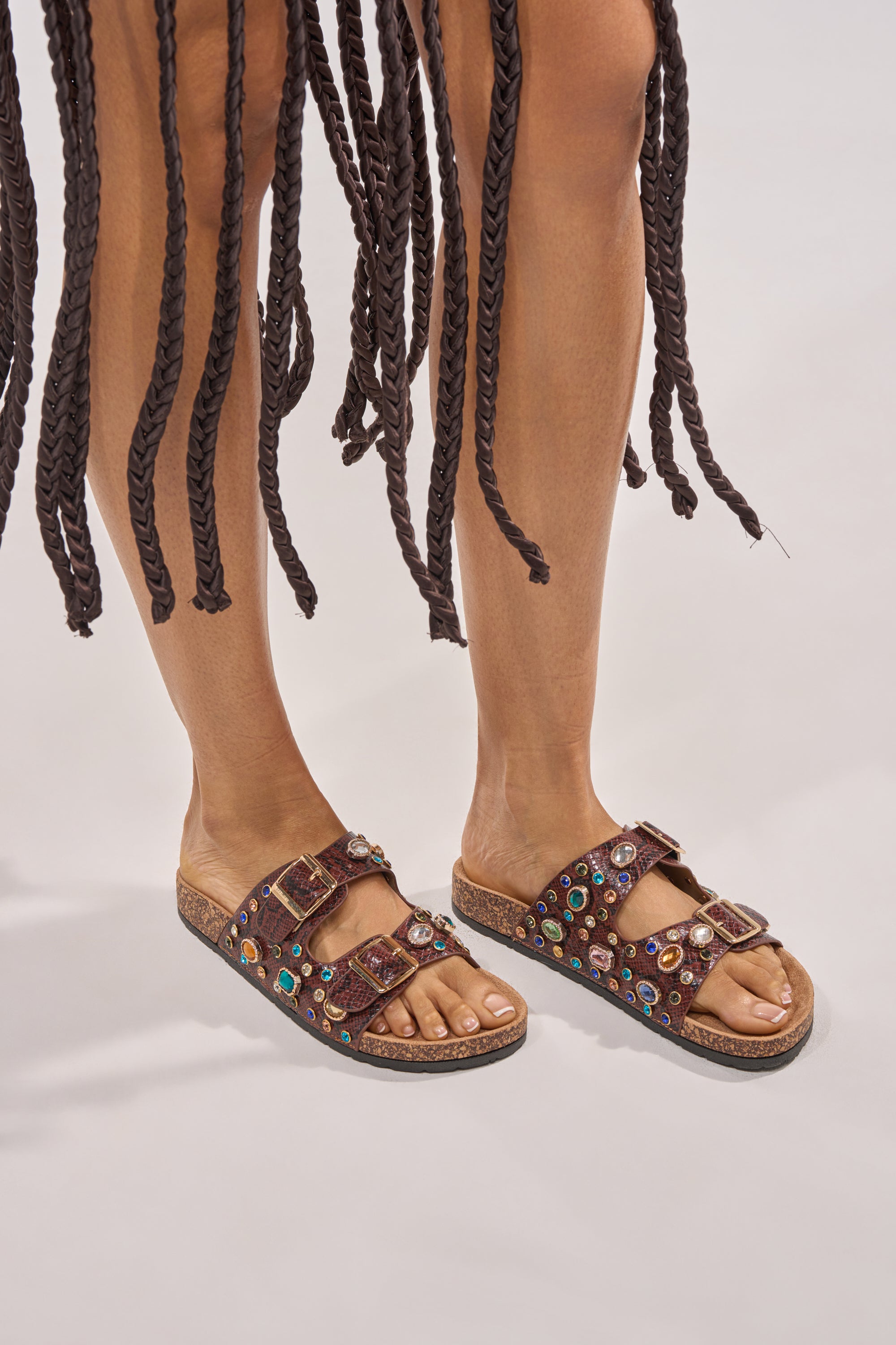 A person wearing AKIRA BAYLEAVES BROWN SNAKE EMBELLISHED FLAT SLIP ON SANDALS with two straps and cork soles stands on a light surface, long thick dark braids from their outfit hanging around their legs. 3