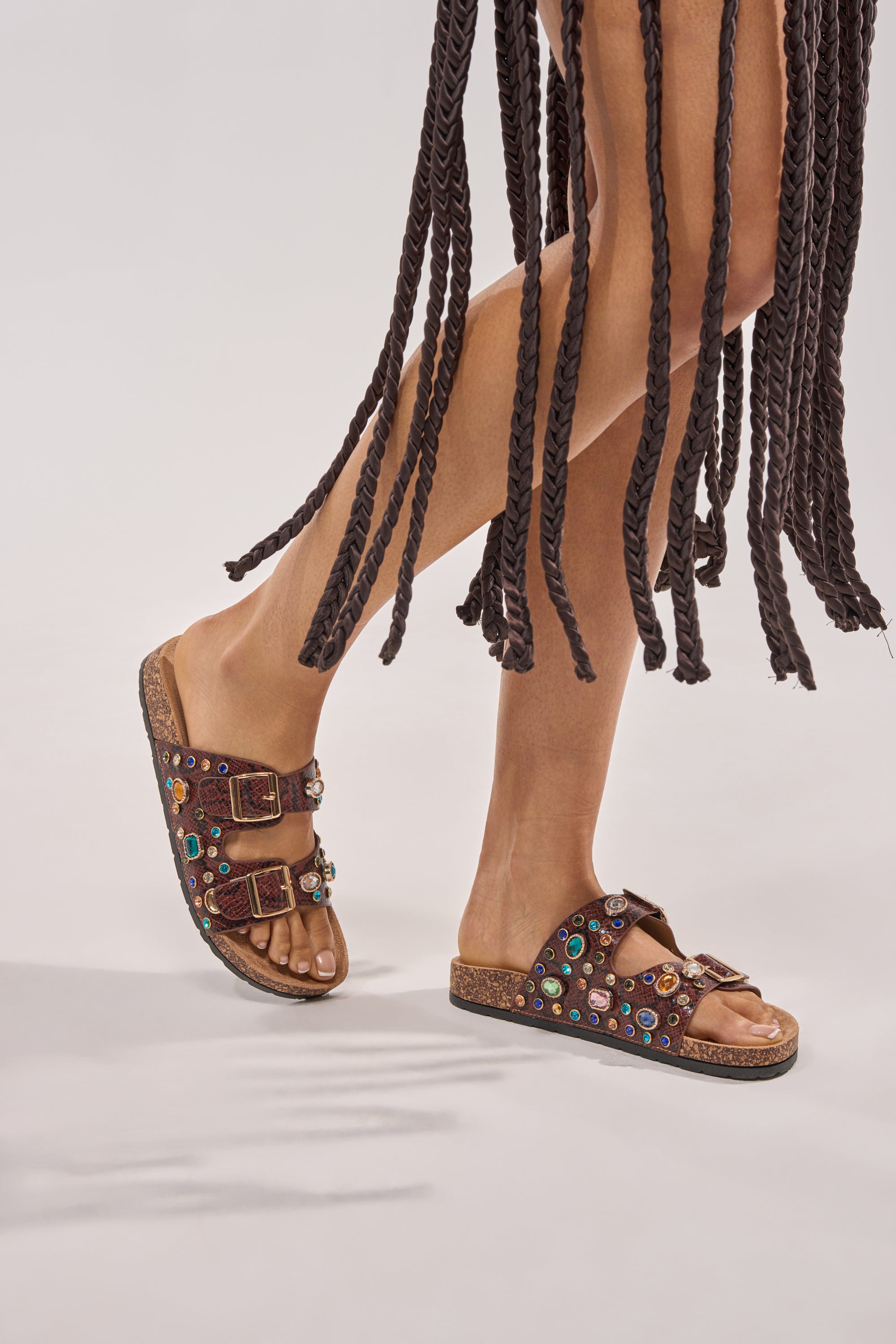 A person stands with one leg crossed in front of the other, wearing a skirt of thick braided strands and AKIRA BAYLEAVES BROWN SNAKE EMBELLISHED FLAT SLIP ON SANDALS; the background is plain and light-colored. 5