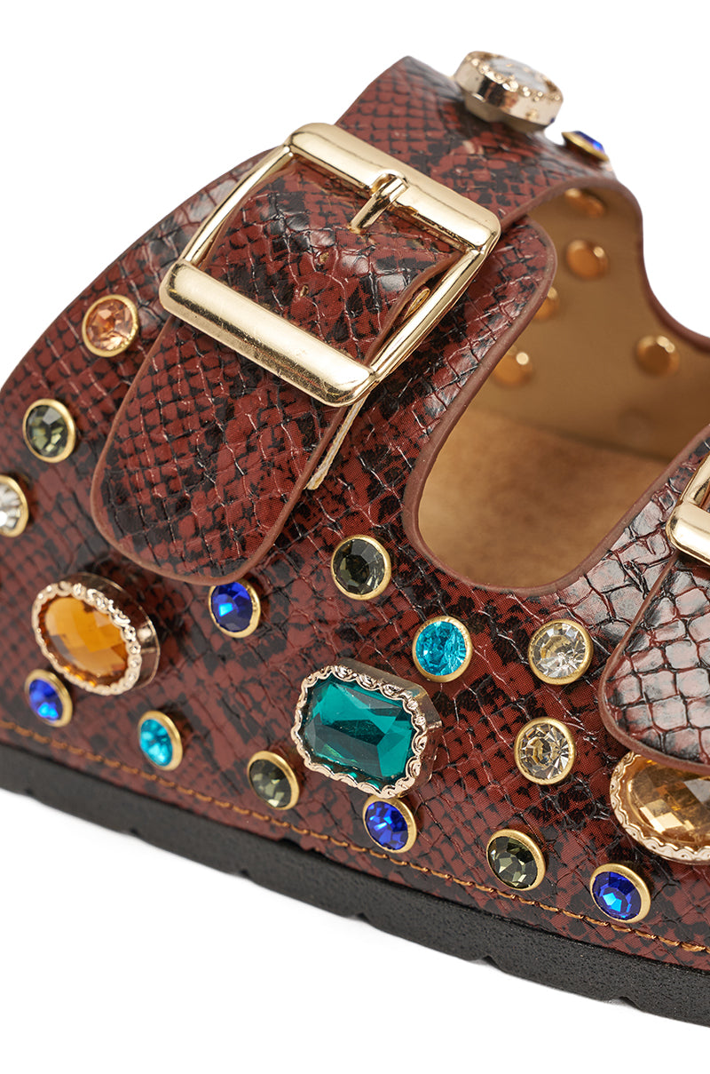 The AKIRA BAYLEAVES BROWN SNAKE EMBELLISHED FLAT SLIP ON SANDAL features a brown snakeskin design, gold buckle, and bold multicolored gemstone studs in assorted shapes adorning the strap and side. 9