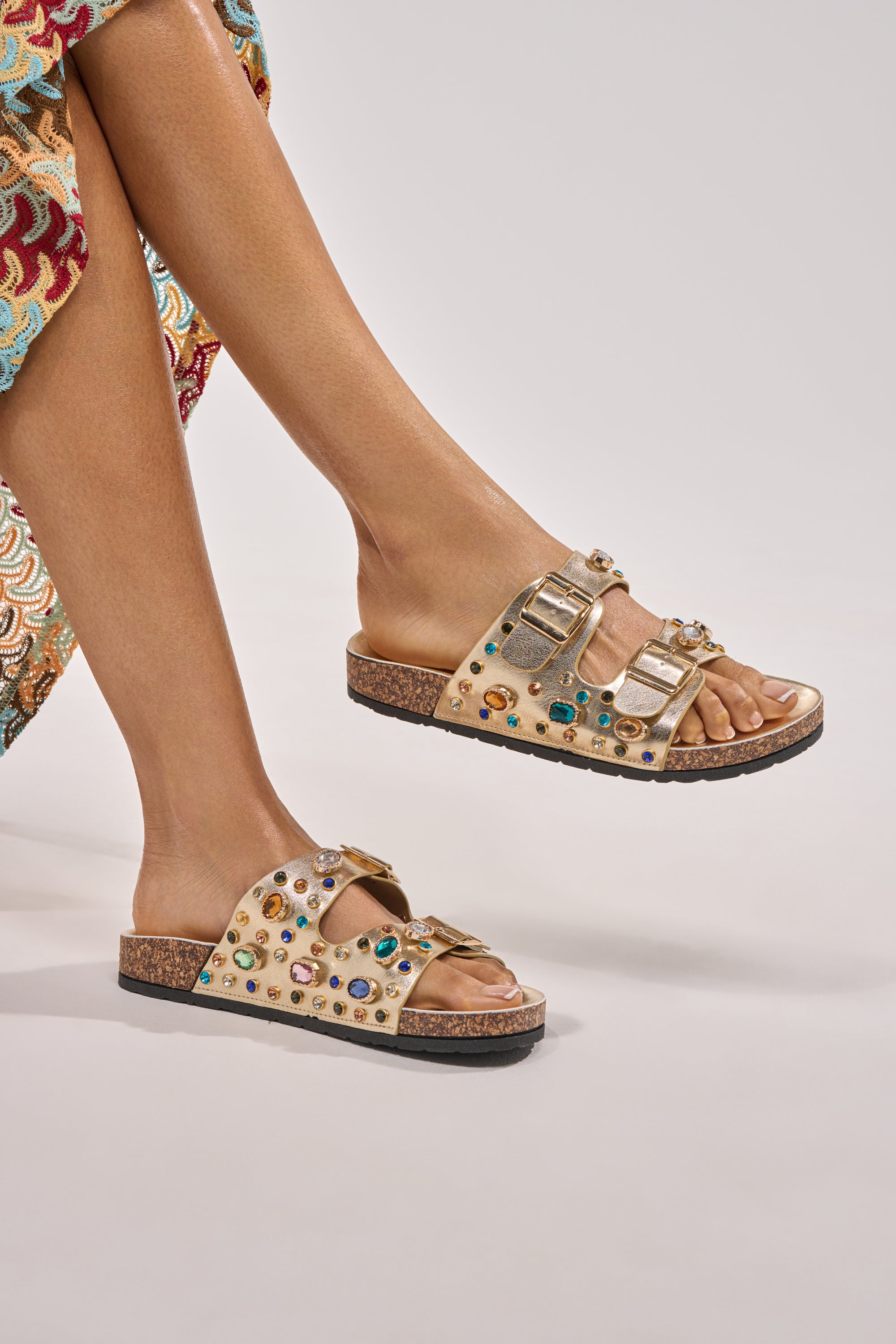 A person wearing a colorful patterned skirt and the AZALEA WANG BAYLEAVES GOLD EMBELLISHED SANDAL, featuring multicolored gems and metallic buckles, sits with legs crossed against a plain light background. 2