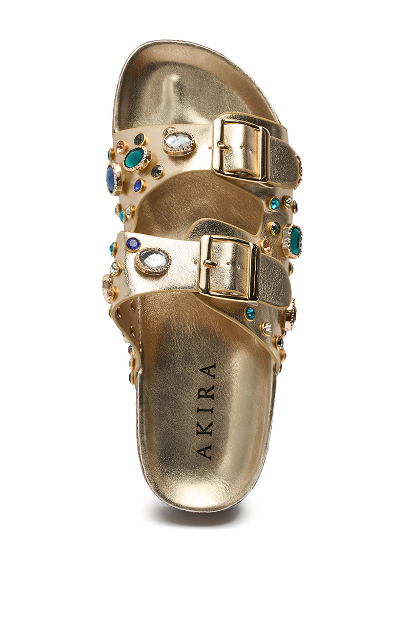 The AZALEA WANG BAYLEAVES GOLD EMBELLISHED SANDAL features a gold faux leather design with two buckle straps adorned with colorful gemstones and rhinestones, plus a metallic gold insole branded "AKIRA." Ideal for adding sparkle to your summer beach look. 8