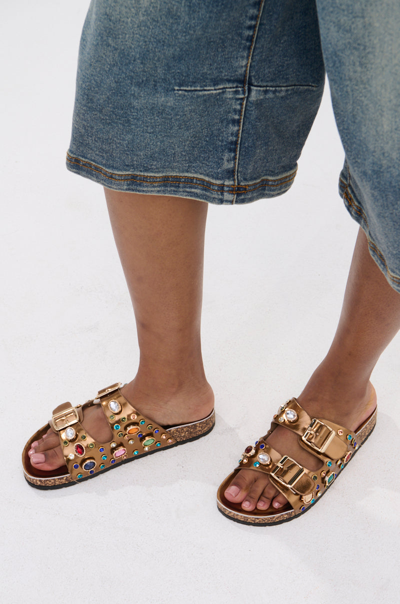 A person wearing denim knee-length shorts and the AKIRA BAYLEAVES BRONZE SLIP ON FLAT SANDAL, featuring faux leather, rhinestone studs, and gold buckles, stands on a light surface.