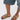 A person in knee-length denim shorts and AKIRA BAYLEAVES GOLD NEW SLIP ON FLAT SANDAL, featuring a faux leather upper and chunky gemstone details, stands on a white surface.
