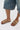 A person in knee-length denim shorts and AKIRA BAYLEAVES GOLD NEW SLIP ON FLAT SANDAL, featuring a faux leather upper and chunky gemstone details, stands on a white surface.