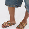 A person in knee-length denim shorts and AKIRA BAYLEAVES GOLD NEW SLIP ON FLAT SANDAL, featuring a faux leather upper and chunky gemstone details, stands on a white surface.