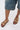 A person wearing knee-length denim shorts and AKIRA BAYLEAVES GOLD NEW SLIP ON FLAT SANDALS with chunky gemstone detailing on a white surface.