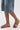 A person in knee-length denim shorts and AKIRA BAYLEAVES GOLD NEW SLIP ON FLAT SANDALS featuring chunky gemstone details and a faux leather upper stands on a light-colored floor.