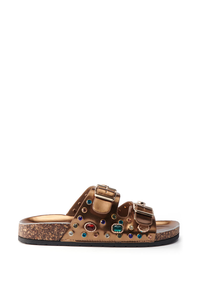 The AKIRA BAYLEAVES BRONZE SLIP ON FLAT SANDAL features two buckled faux leather straps with rhinestone studs and colorful gemstones on a cork-textured sole, shown against a white background.