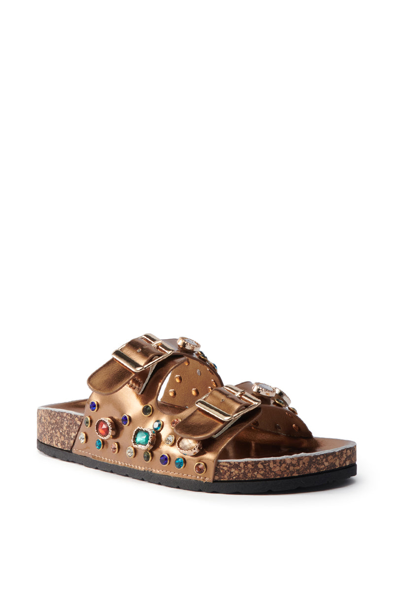 The AKIRA BAYLEAVES BRONZE SLIP ON FLAT SANDAL features two large buckled faux leather straps with rhinestone studs and colorful gemstones, plus a cork sole and black rubber bottom, shown on a white background.