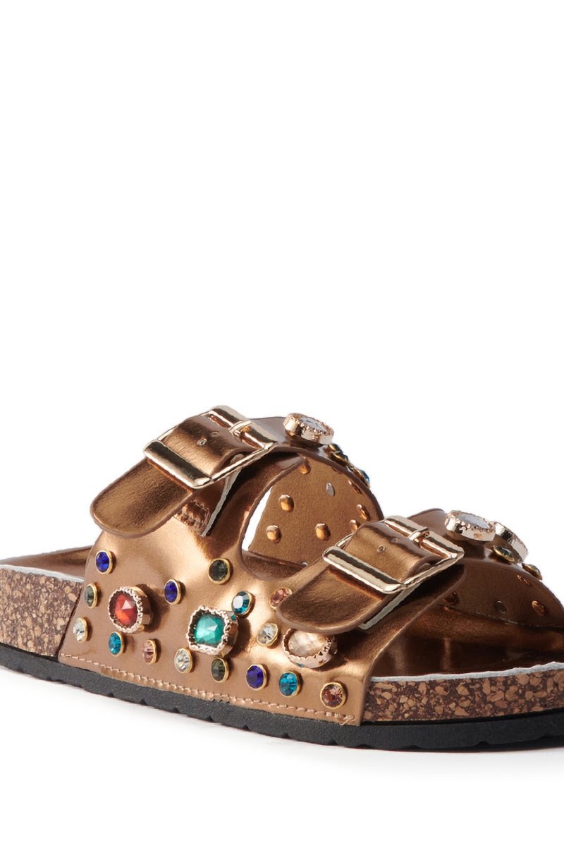 The AKIRA BAYLEAVES BRONZE SLIP ON FLAT SANDAL features bronze faux leather, two large buckle straps with colorful rhinestone studs, and a cork sole with a black bottom.