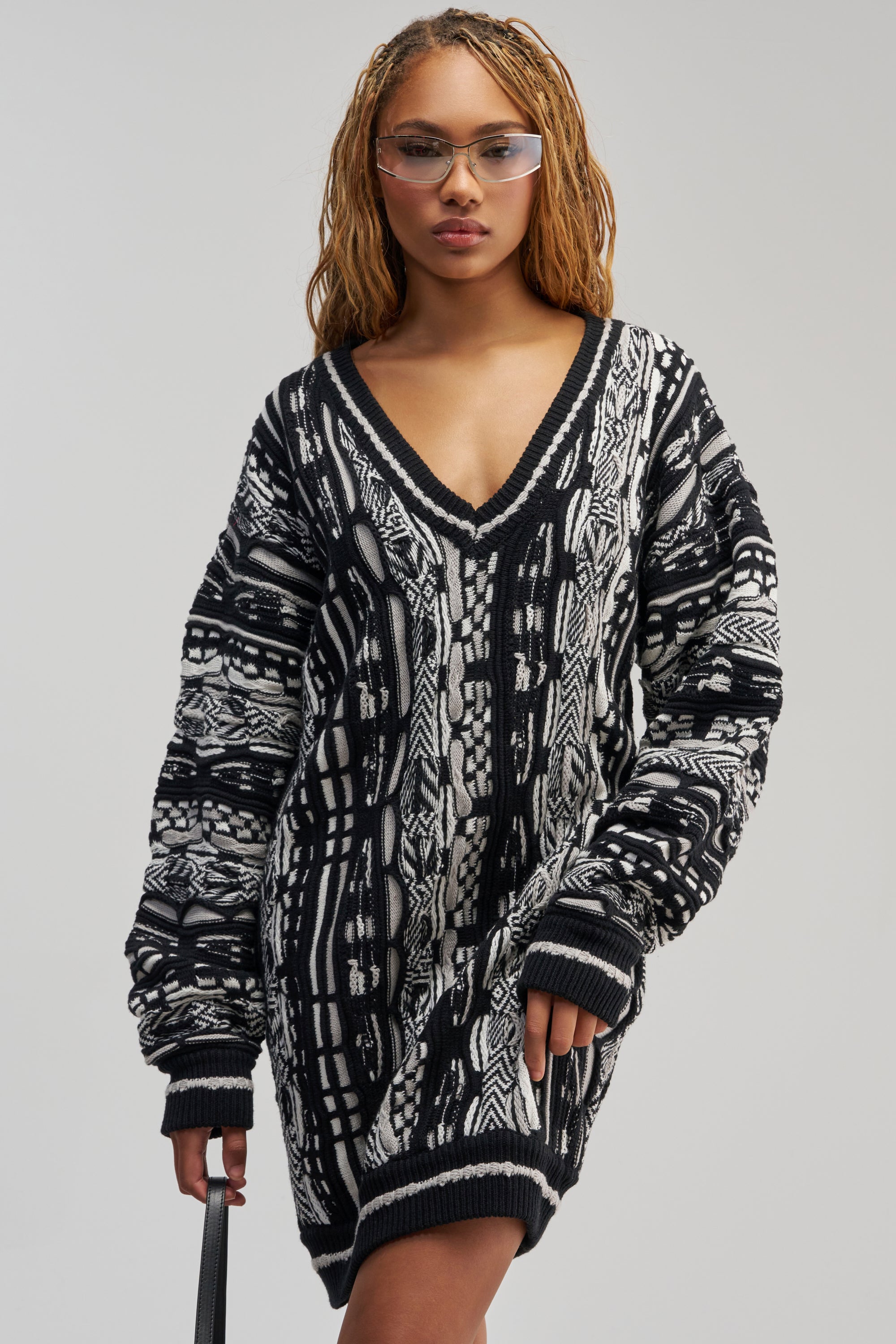 A person wearing the FEELIN COZY V-NECK SWEATER DRESS IN BLACK AND WHITE and clear glasses stands against a plain background, holding a black strap in their left hand.