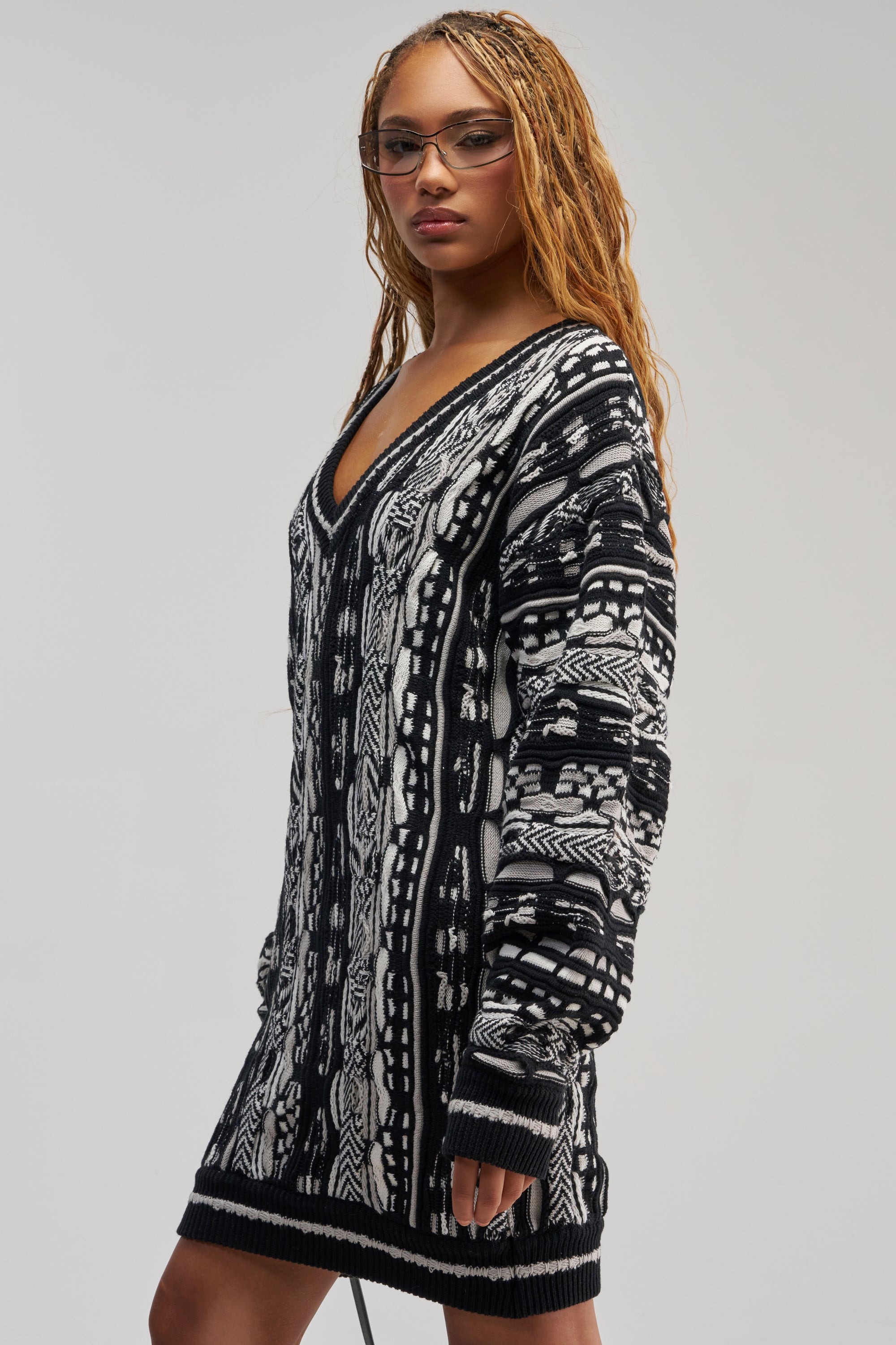 A person with long braided hair and glasses models the FEELIN COZY V-NECK SWEATER DRESS IN BLACK AND WHITE, posing confidently against a plain gray background.