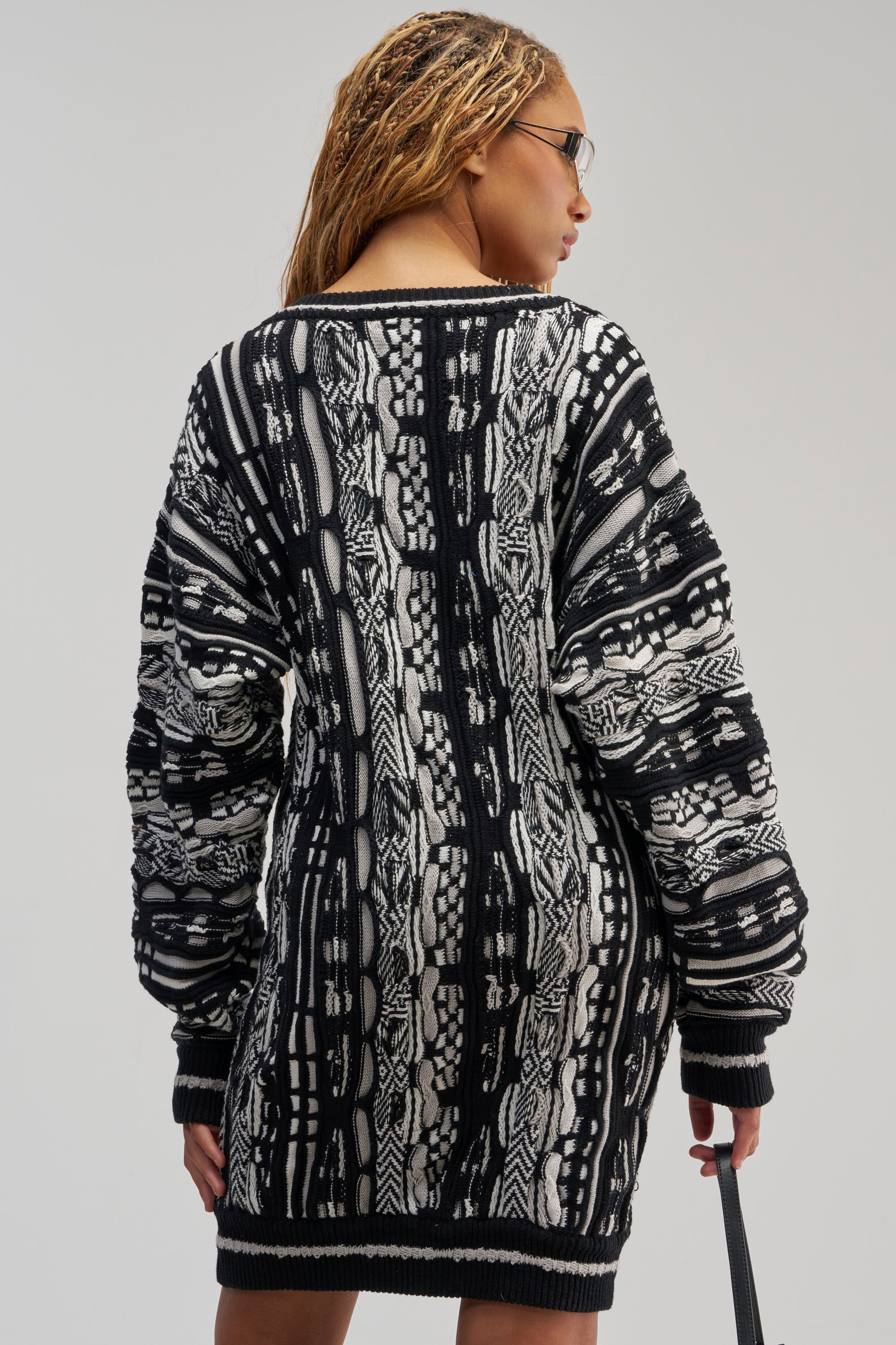 A person with braided hair, wearing glasses and the FEELIN COZY V-NECK SWEATER DRESS IN BLACK AND WHITE—a long-sleeve knit mini dress—is shown from the back against a plain background.