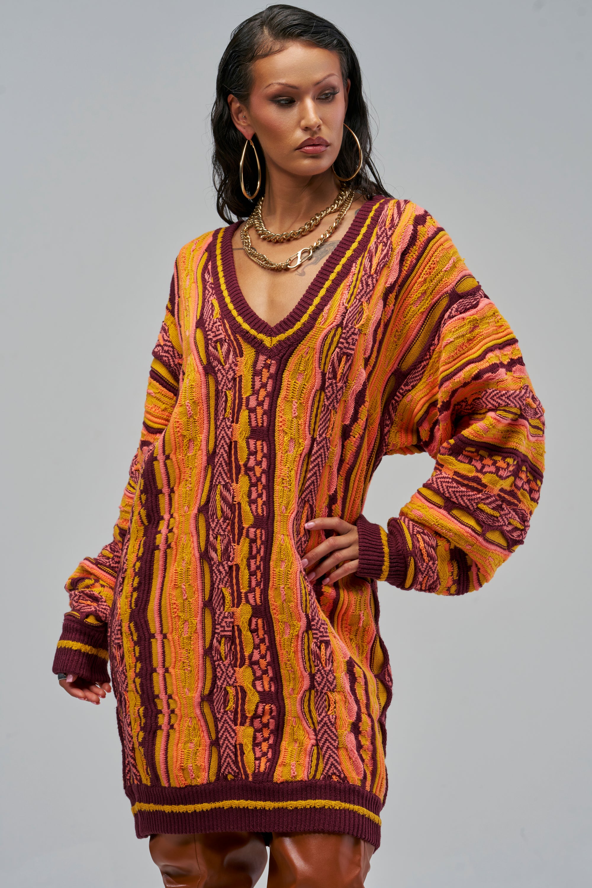 A model wearing the FEELIN COZY V-NECK SWEATER DRESS IN ORANGE, an oversized, long-sleeved textured knit mini dress featuring a vertical, multicolored pattern in shades of orange, yellow, and burgundy.
