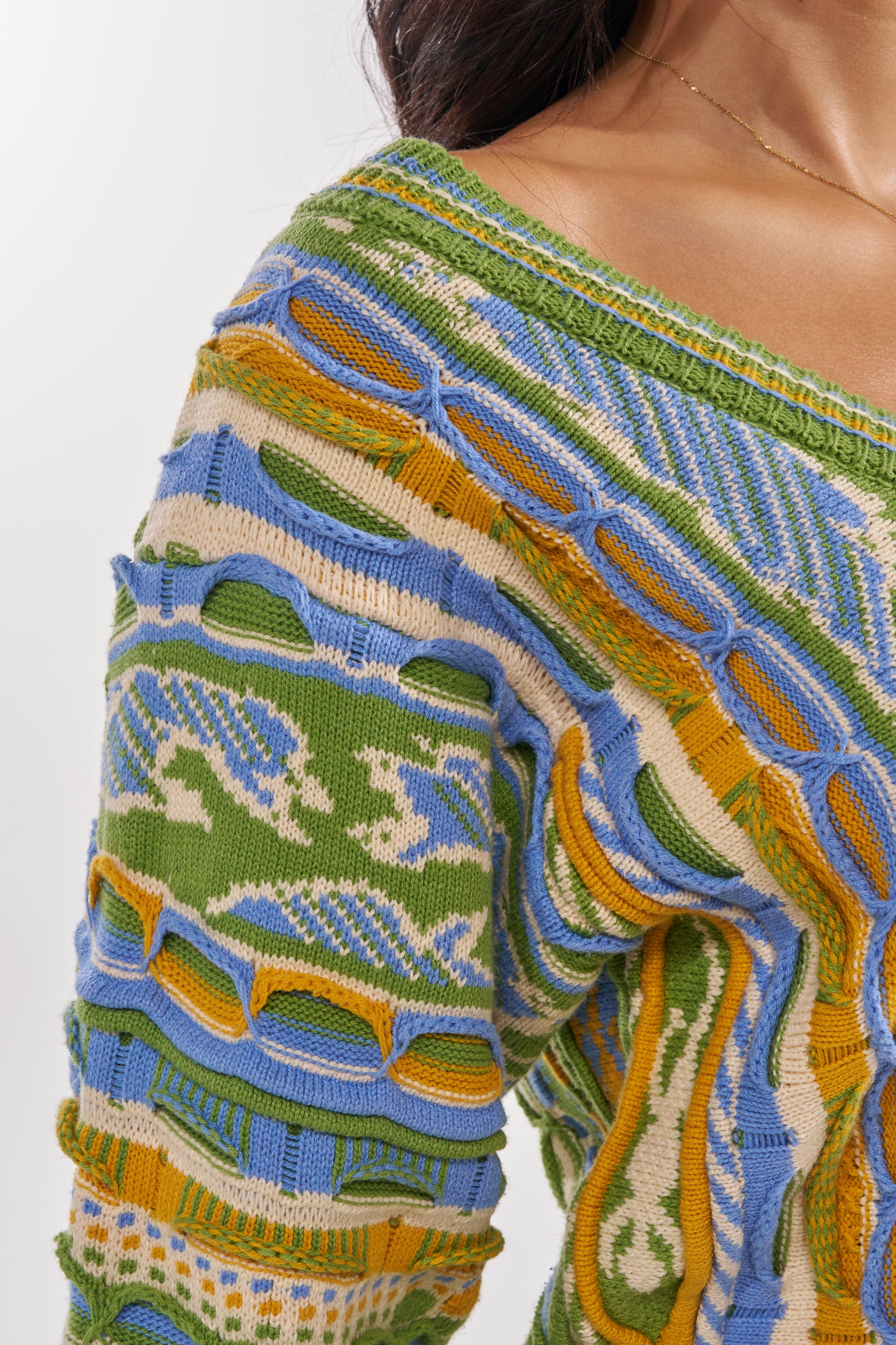 A close-up of a person wearing the FEELIN COZY V-NECK SWEATER DRESS, a colorful knit featuring green, blue, yellow, and cream patterns with raised wavy details. The intricate multicolor design is shown; the person's face is not visible. 6