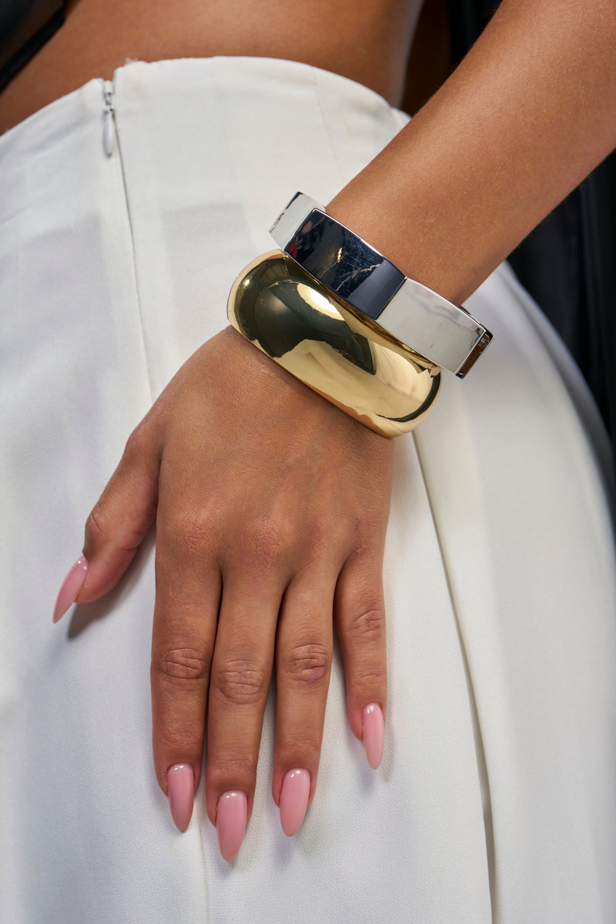 A hand with long, pink nails rests on white pants. The wrist is adorned with the TWO TONE METAL BANGLE SET, a chunky gold and silver accessory featuring an elegant hinge closure for easy wear.