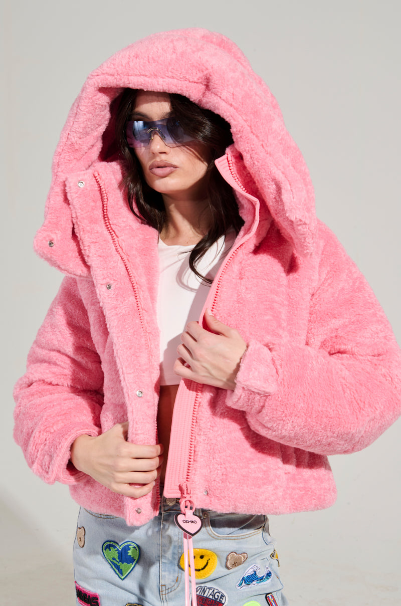 Wearing the MARDIE Pink Fuzzy Hooded Puffer with an adjustable cinched waist and a pair of blue sunglasses, a person stands casually. They are paired with light-colored jeans featuring colorful patches against a plain and minimalistic background.