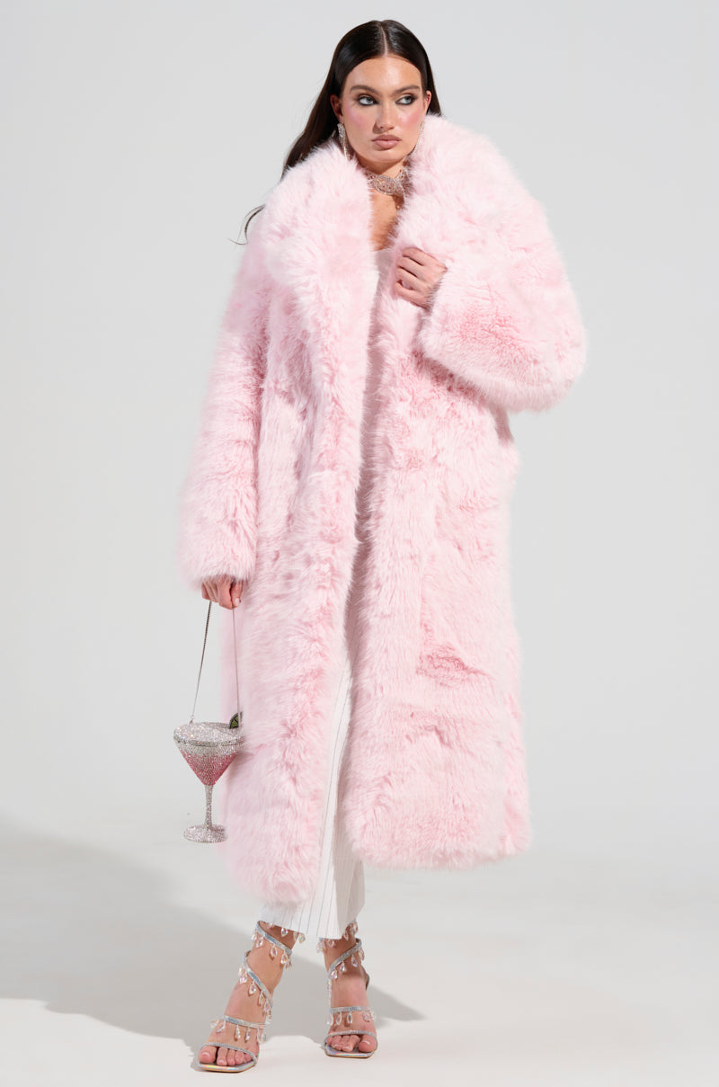 Dressed in the BEAR HUG LUXE FAUX FUR TRENCH COAT IN PINK, a woman elegantly holds a large, decorative cocktail glass. She pairs this luxurious coat with jeweled sandals and a sleek white outfit underneath, standing against a plain background.