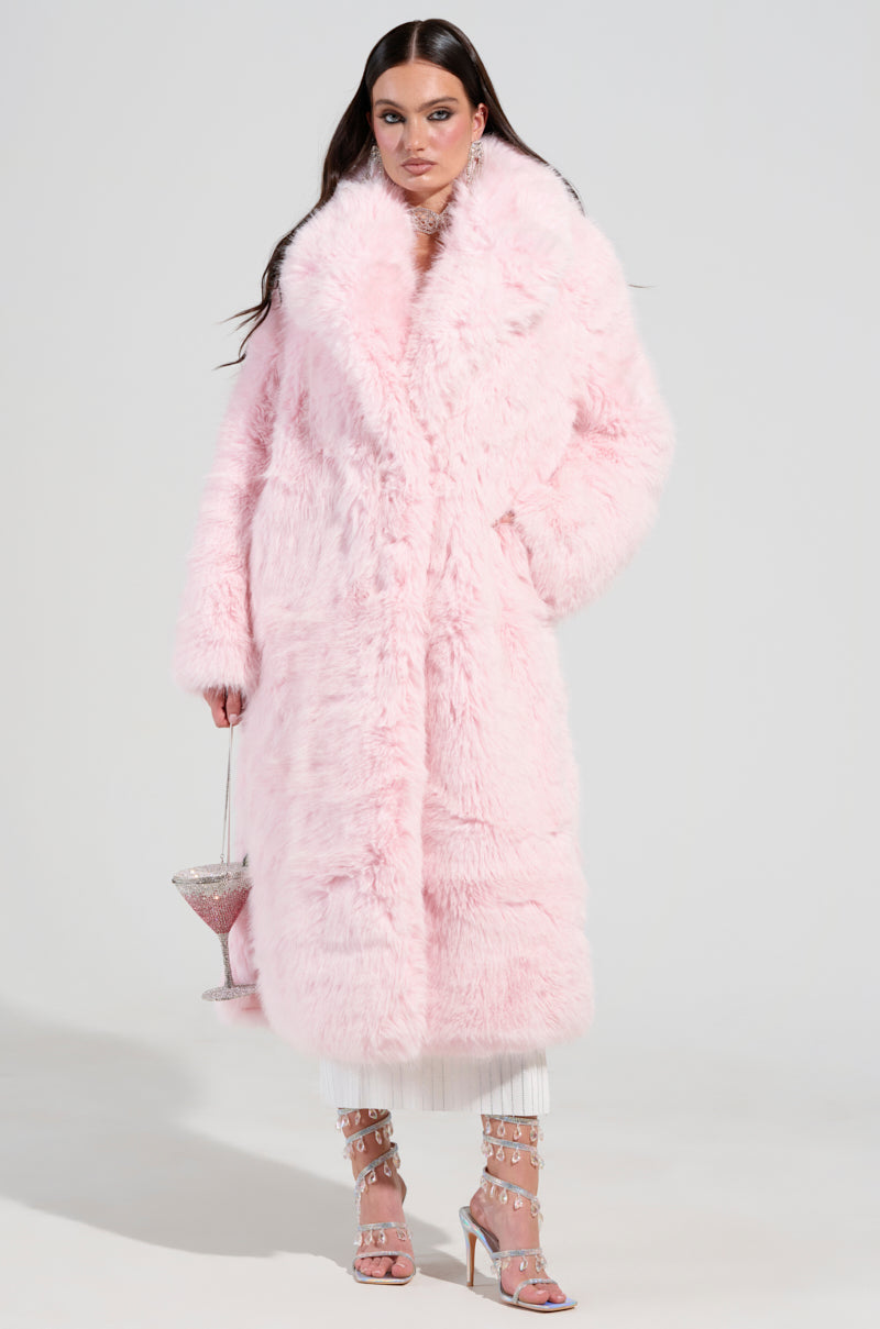 A person poses in the elegant BEAR HUG LUXE FAUX FUR TRENCH COAT IN PINK, holding a small purse. They are dressed in white pants and pink heels embellished with rhinestones, all set against a plain white studio backdrop.