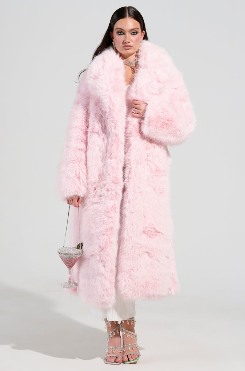A woman stands confidently in the luxurious BEAR HUG LUXE FAUX FUR TRENCH COAT IN PINK, holding an oversized crystal goblet filled with a pink drink. She pairs it with white pants and embellished high-heeled sandals, all set against a plain white background.