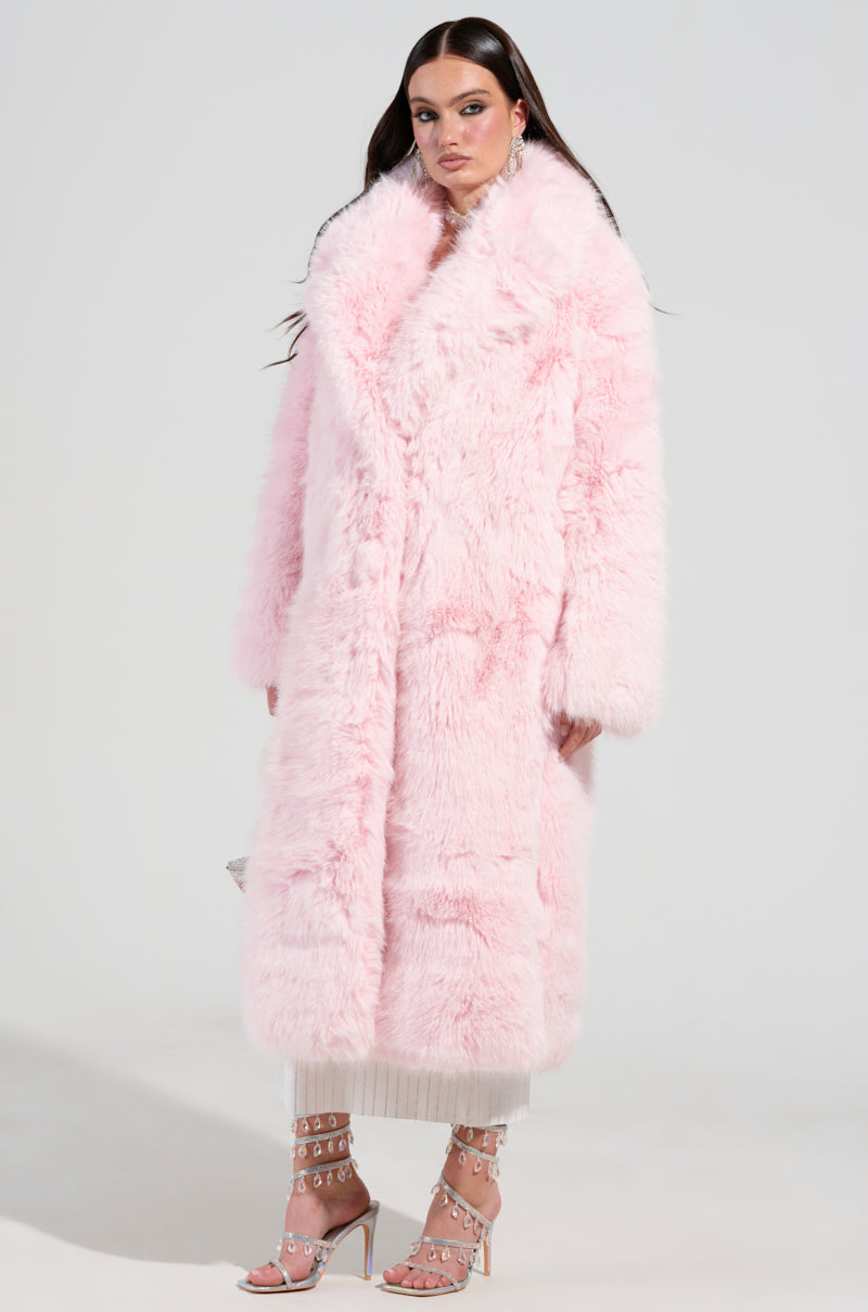 Wearing the BEAR HUG LUXE FAUX FUR TRENCH COAT in pink, a woman stands confidently, exuding elegance. Her look is completed with strappy high heels and a long skirt. With her long, dark hair cascading down, she poses against a plain, light background, showcasing timeless sophistication.