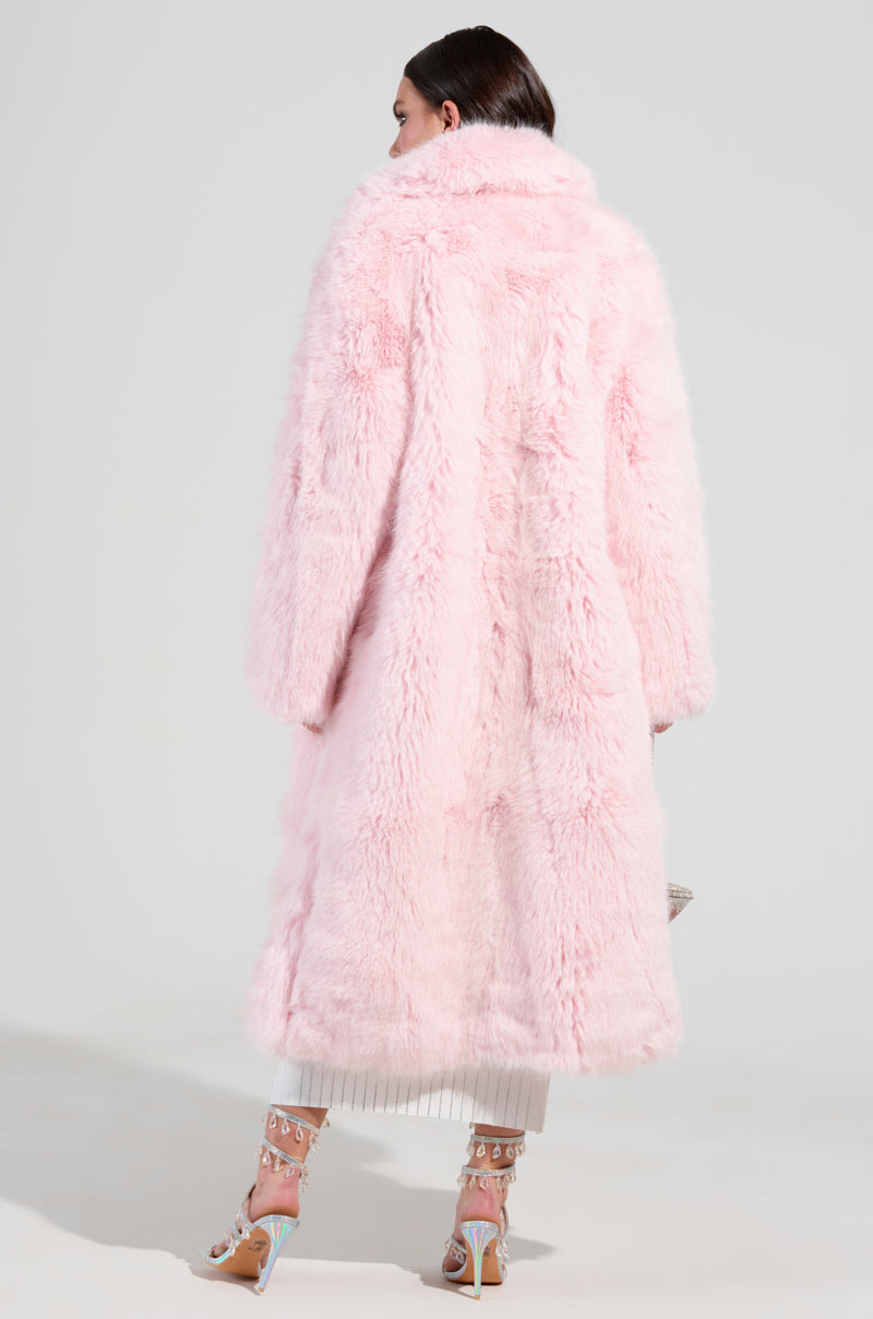 A person wearing the BEAR HUG LUXE FAUX FUR TRENCH COAT IN PINK stands against a plain background, their back turned to the camera. Stylish high-heeled sandals with embellishments complement the look, along with a pleated white skirt peeking from beneath the long, fluffy pink ensemble.