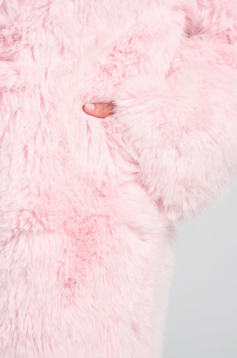 Close-up of a person wearing the BEAR HUG LUXE FAUX FUR TRENCH COAT IN PINK. The partially visible hand, tucked into a side pocket, highlights the texture and softness of the coat against a plain white background.