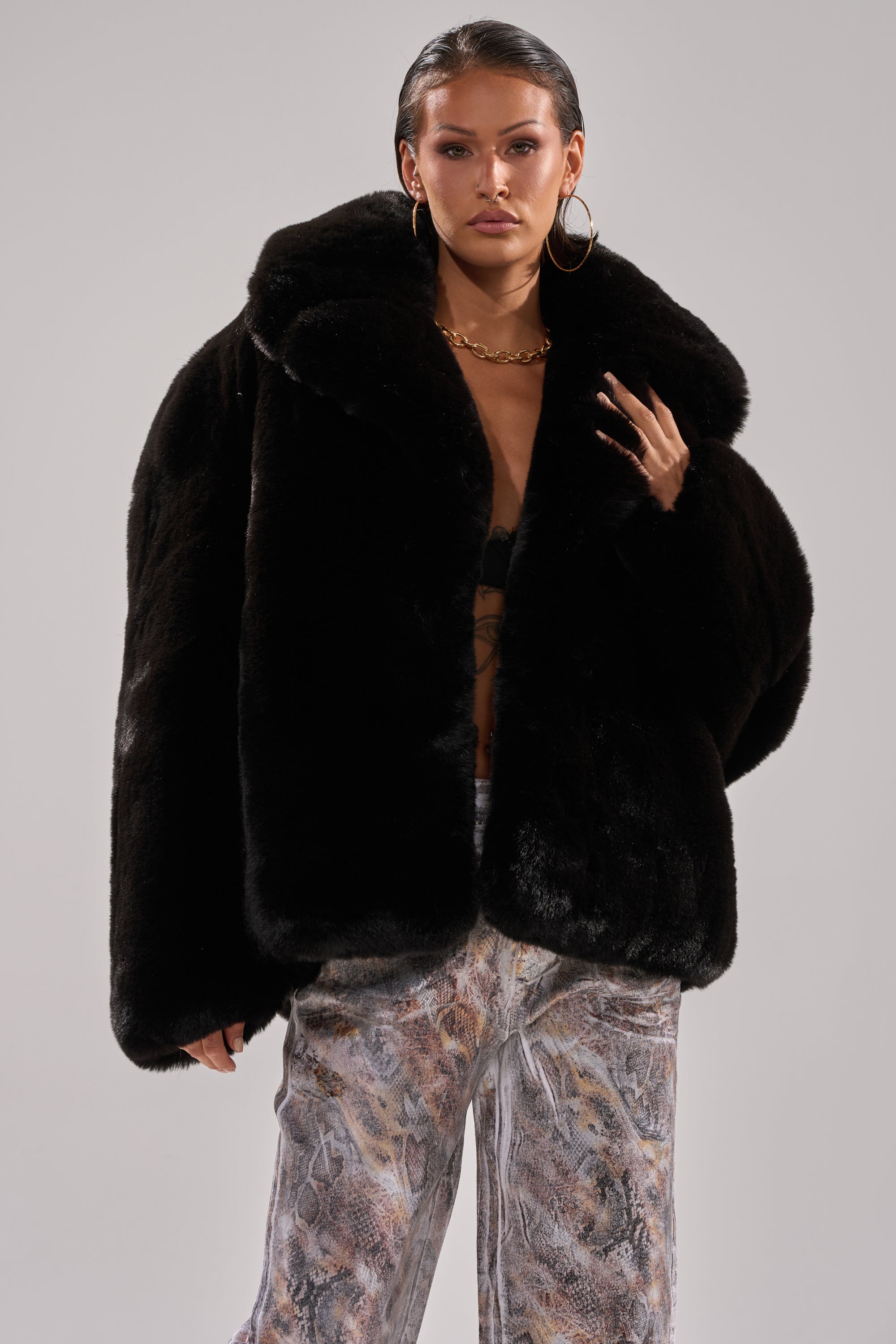 Wearing the NO LOOKING BACK FAUX FUR IN BLACK, hoop earrings, and a gold chain necklace, a woman with slicked-back hair and patterned pants stands confidently against a plain light background.
