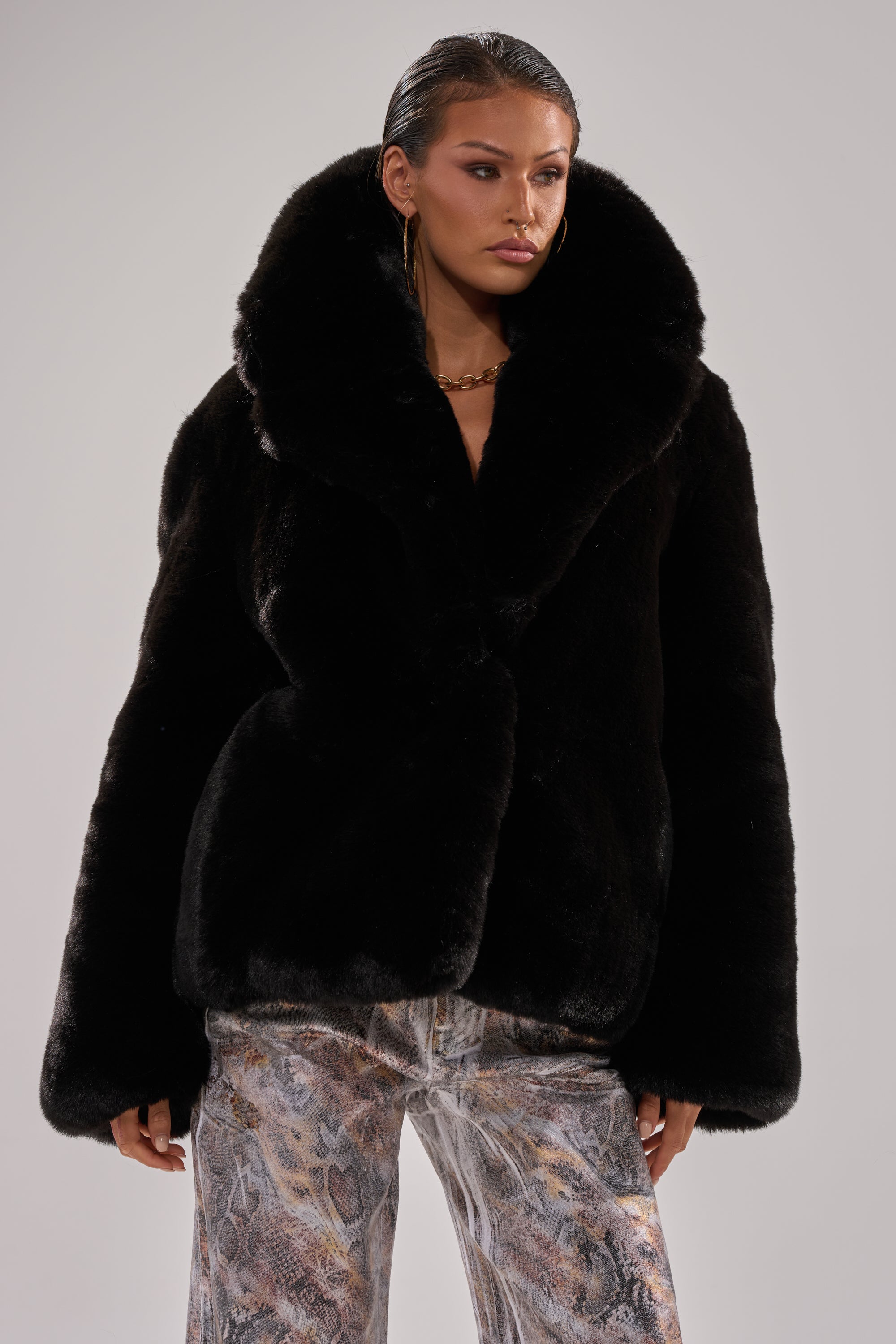 A woman stands against a plain light background, looking to the side in a stylish NO LOOKING BACK FAUX FUR IN BLACK coat with a large collar, paired with patterned metallic pants and a neutral expression.