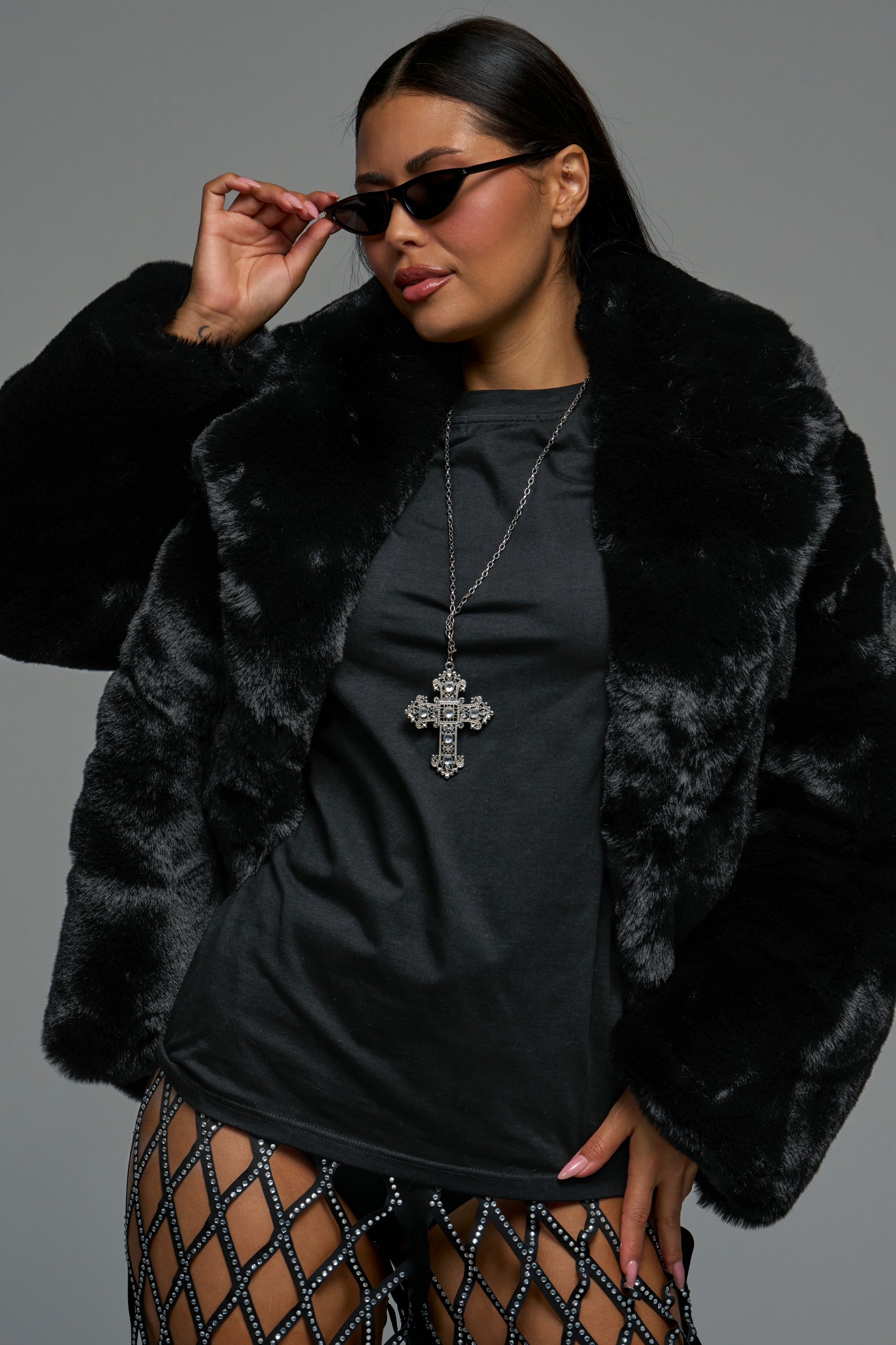 A woman poses confidently against a gray background wearing the NO LOOKING BACK FAUX FUR IN BLACK, paired with black mesh pants, a large cross necklace, and dark sunglasses.