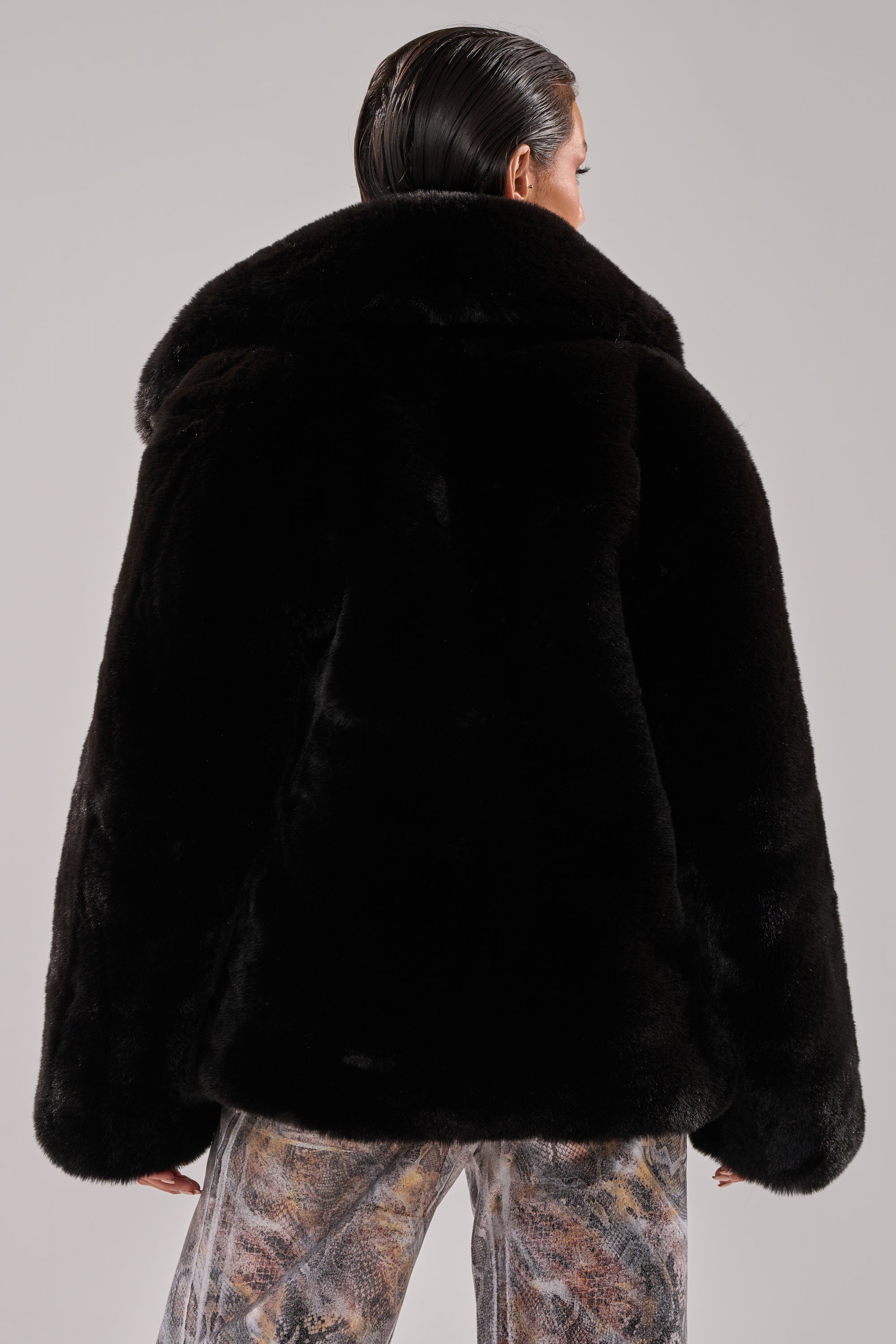 A person with slicked-back hair is shown from behind wearing the NO LOOKING BACK FAUX FUR IN BLACK and patterned pants against a light gray background.