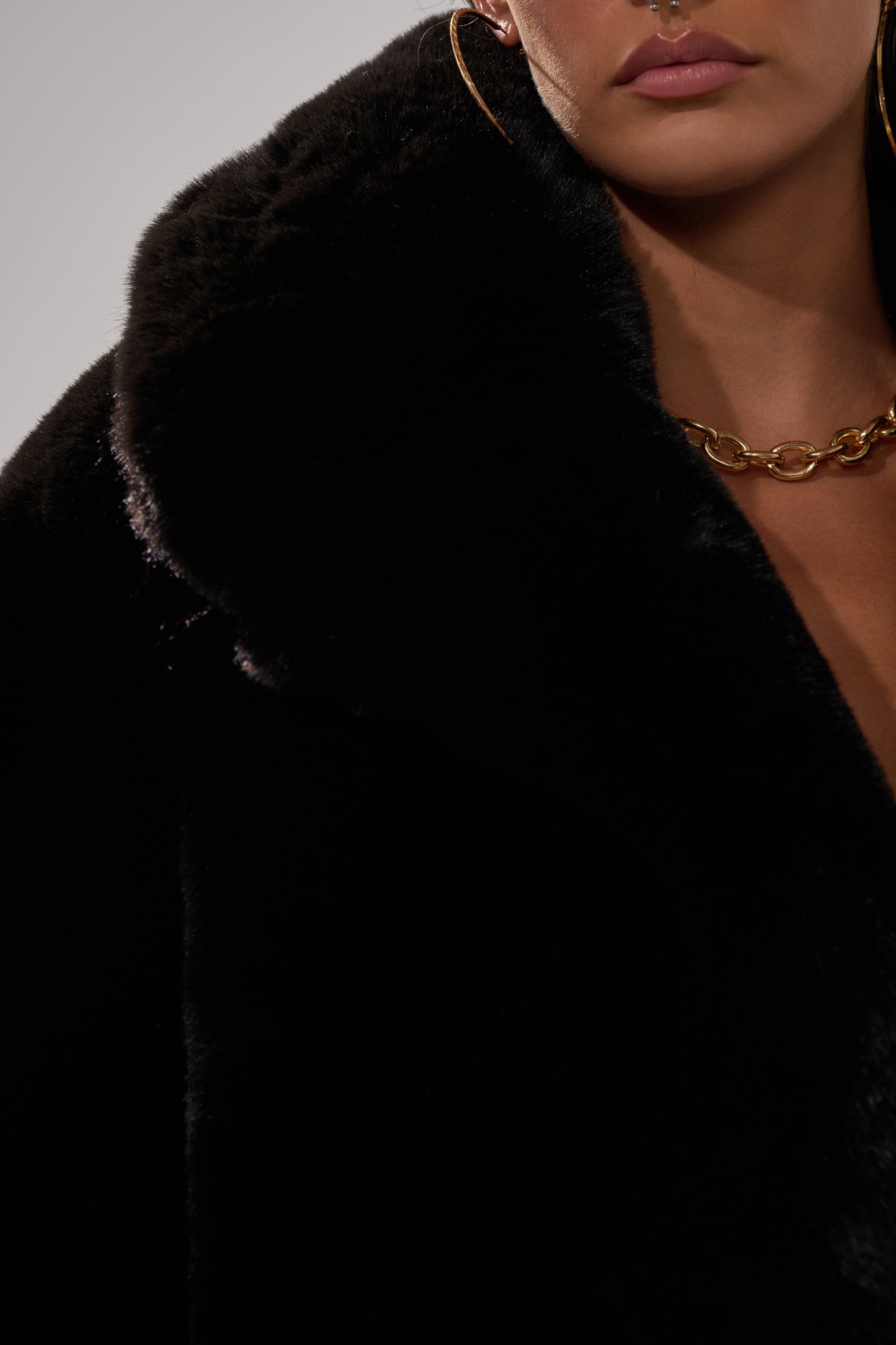 A close-up of a person wearing the NO LOOKING BACK FAUX FUR IN BLACK coat, gold hoop earrings, and a chunky gold chain necklace, showing only the lower half of the face and upper body.
