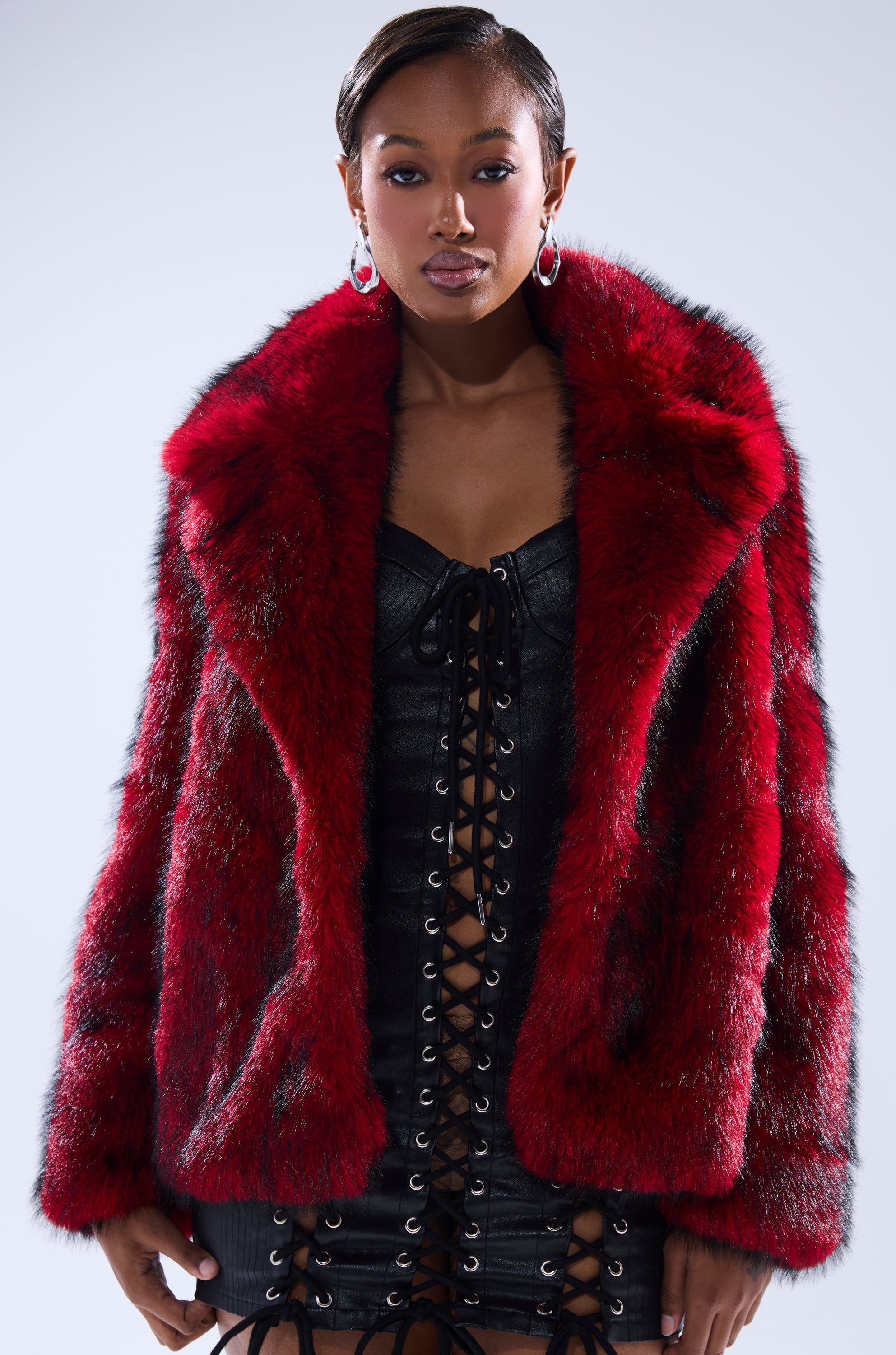 A woman models the NO LOOKING BACK FAUX FUR IN RED MULTI, styled over a black lace-up leather dress, standing confidently against a plain light background and looking directly at the camera.
