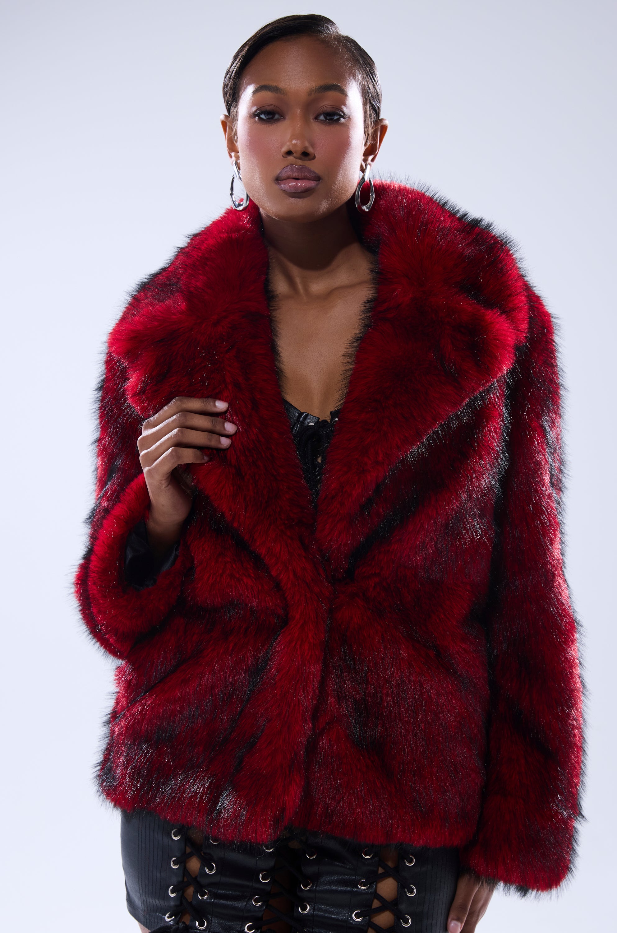 A woman poses confidently in the NO LOOKING BACK FAUX FUR IN RED MULTI, an oversized jacket with bold red and black accents, styled with hoop earrings and a black lace-up outfit against a plain light background.